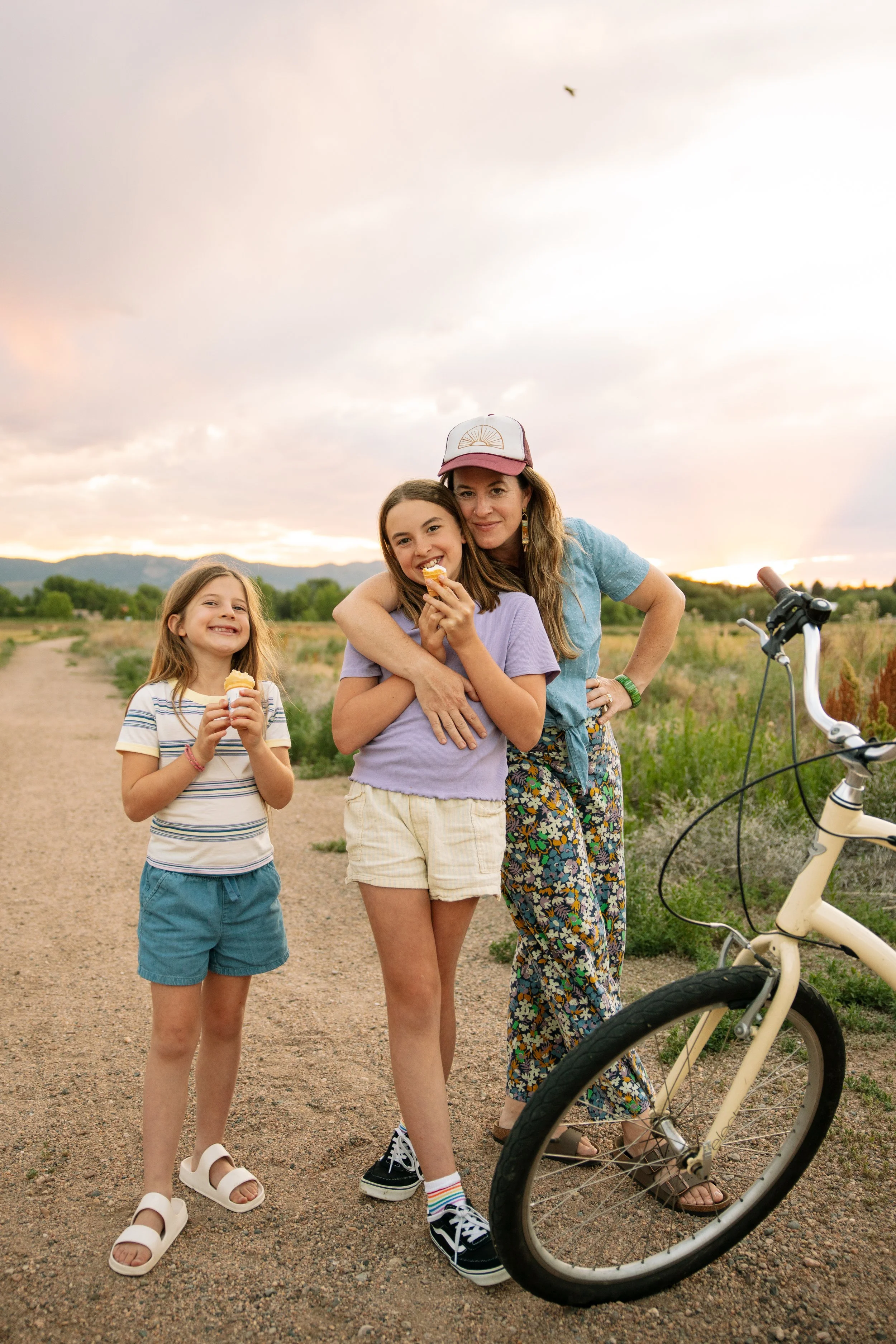 A woman with two young girls standing on a dirt trail outdoors at sunset, holding ice cream cones, with a bicycle nearby and scenic mountains and sky in the background.