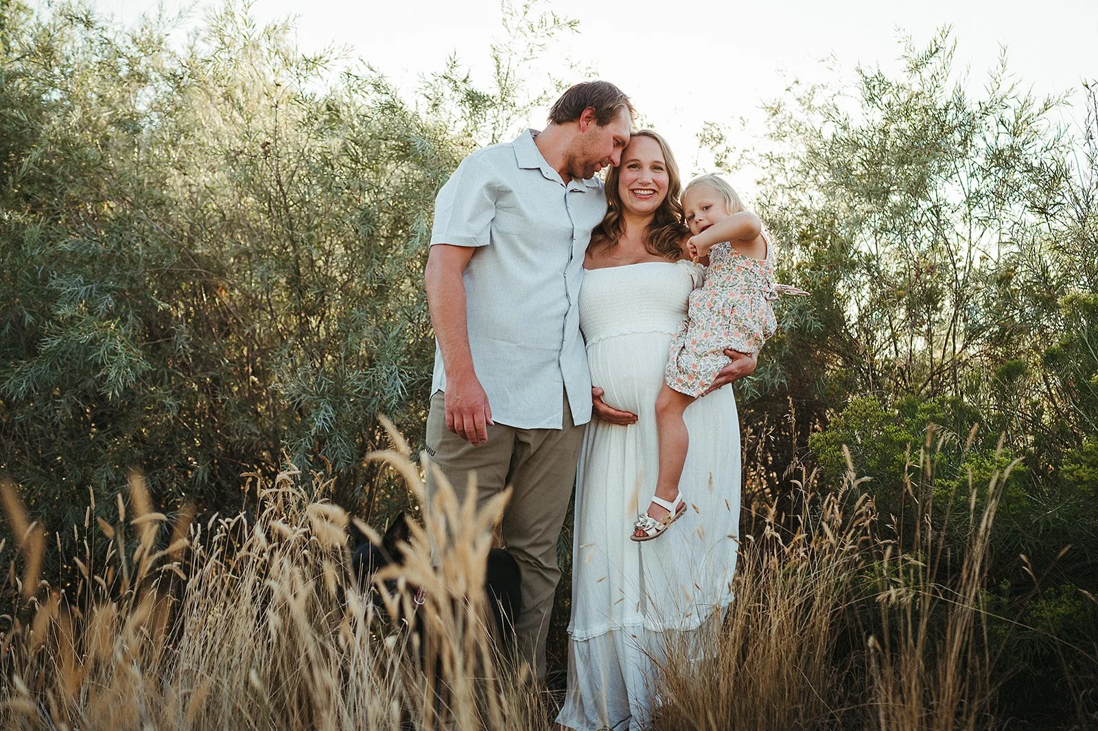 A smiling pregnant woman in a white dress holding a young girl, with a man, standing in a field of tall grass and bushes during sunset.