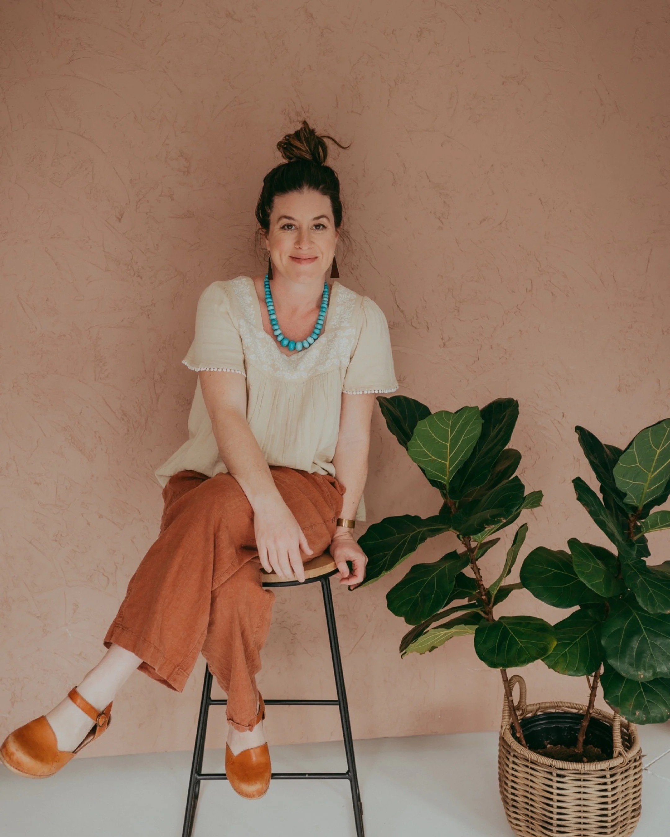 A woman with dark hair in a bun, wearing a cream blouse, brown pants, and tan shoes, sitting on a black stool against a pink textured wall. There is a potted plant to her right with large green leaves, placed in a woven basket.