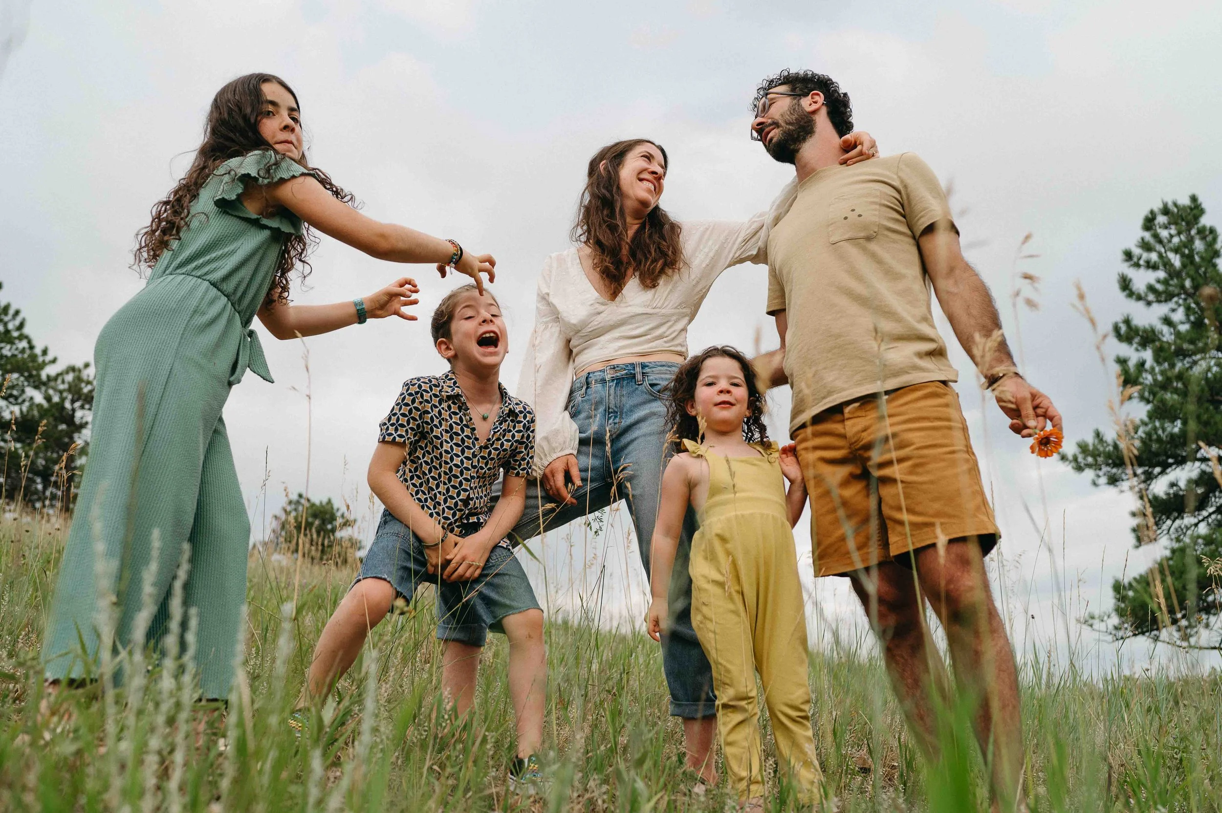 Family of six people standing in a grassy field during daytime, with trees and a cloudy sky in the background. They are smiling and holding hands, appearing happy and enjoying each other's company.