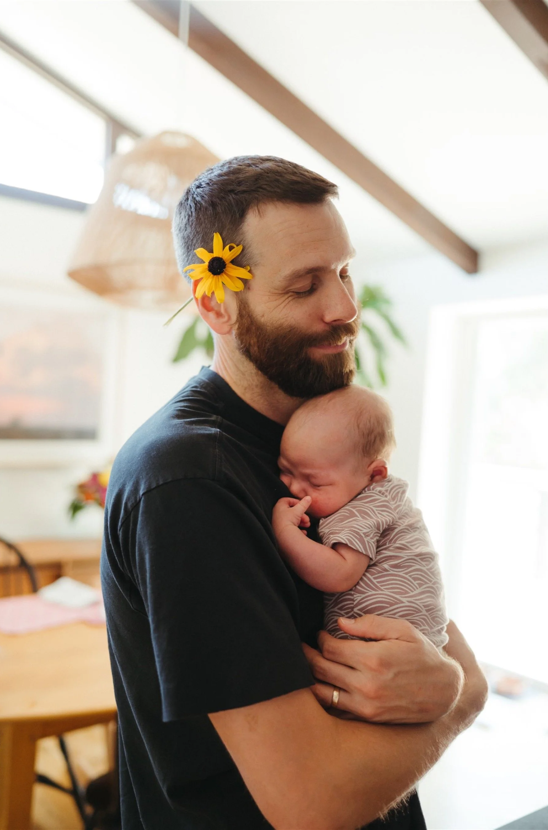 A man with a flower behind his ear hugging a child in a cozy home interior.