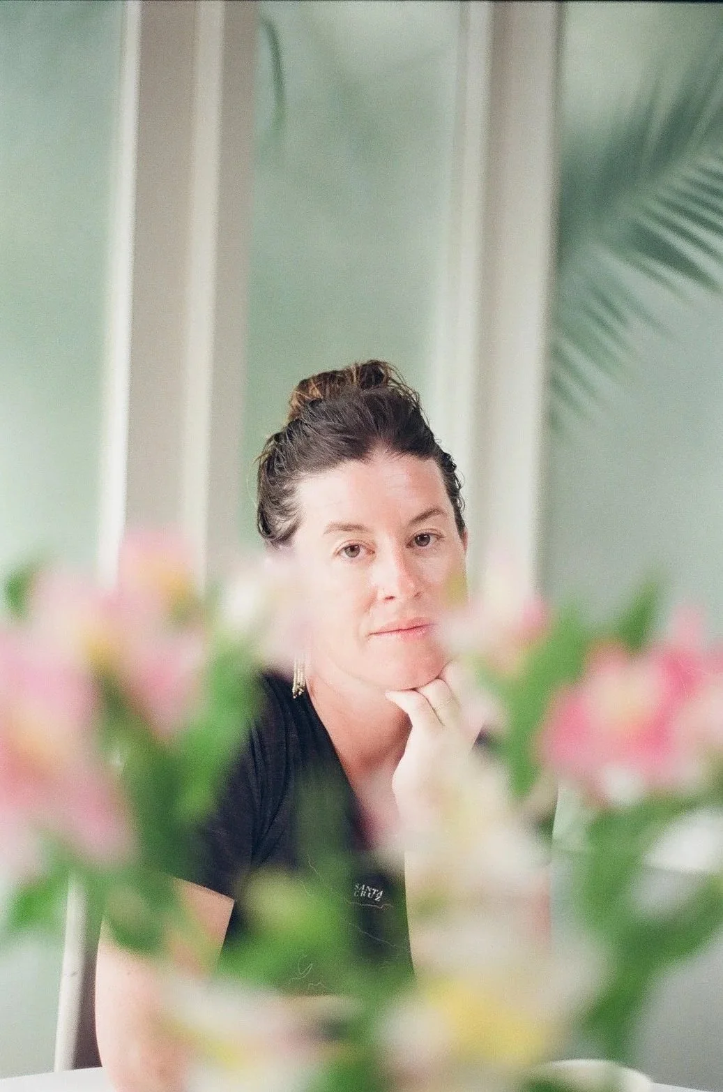 A woman with dark hair pulled back, resting her chin on her hand, looking at the camera through a reflective surface, with pink flowers in the foreground.