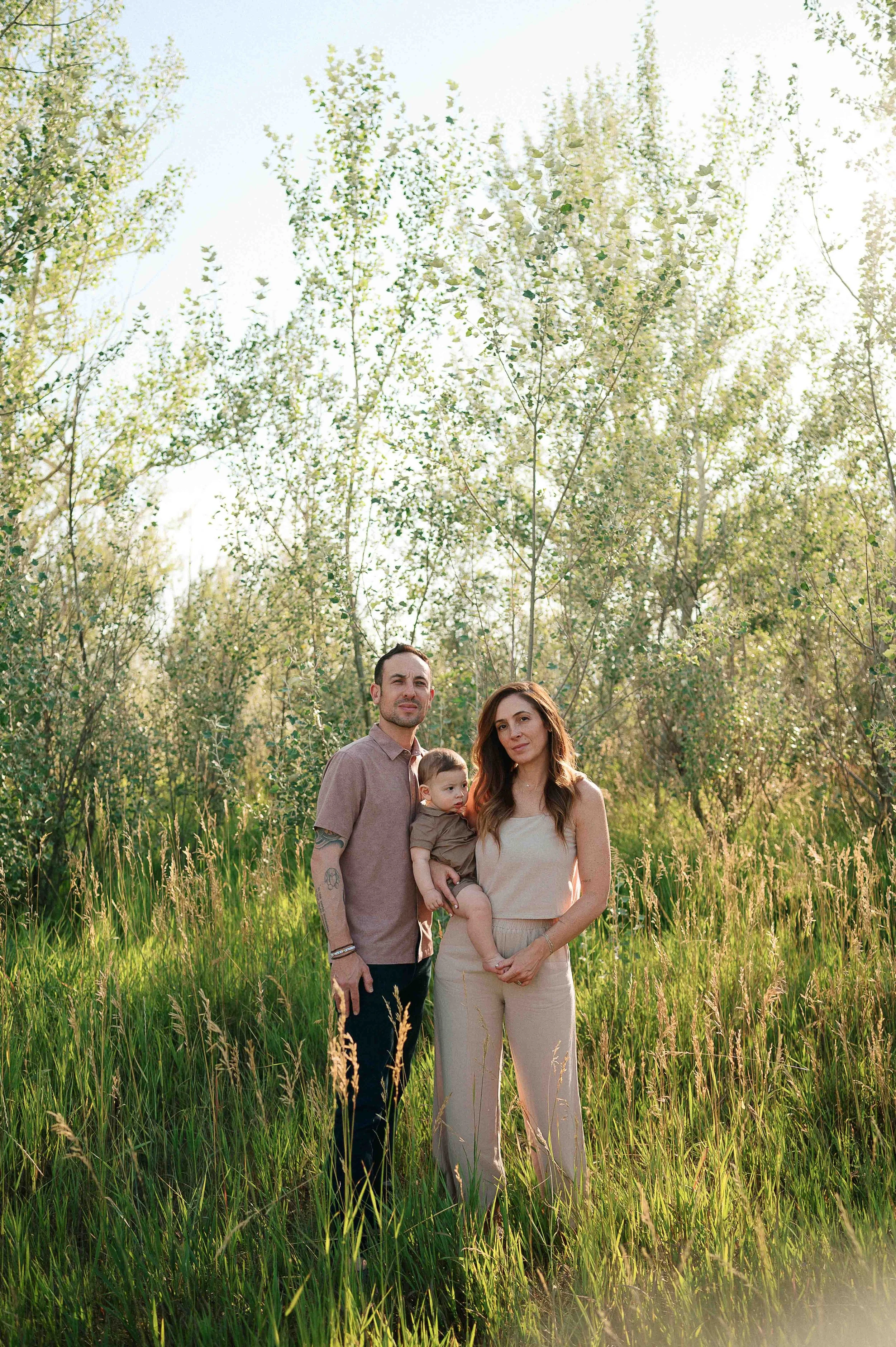 A family of three standing in a grassy field surrounded by trees in bloom, during a sunny day.