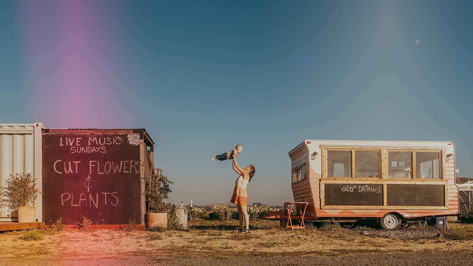 A woman and a child playing outside near a food truck and a sign promoting live music and flower and plant sales, with a clear sky and the moon visible.