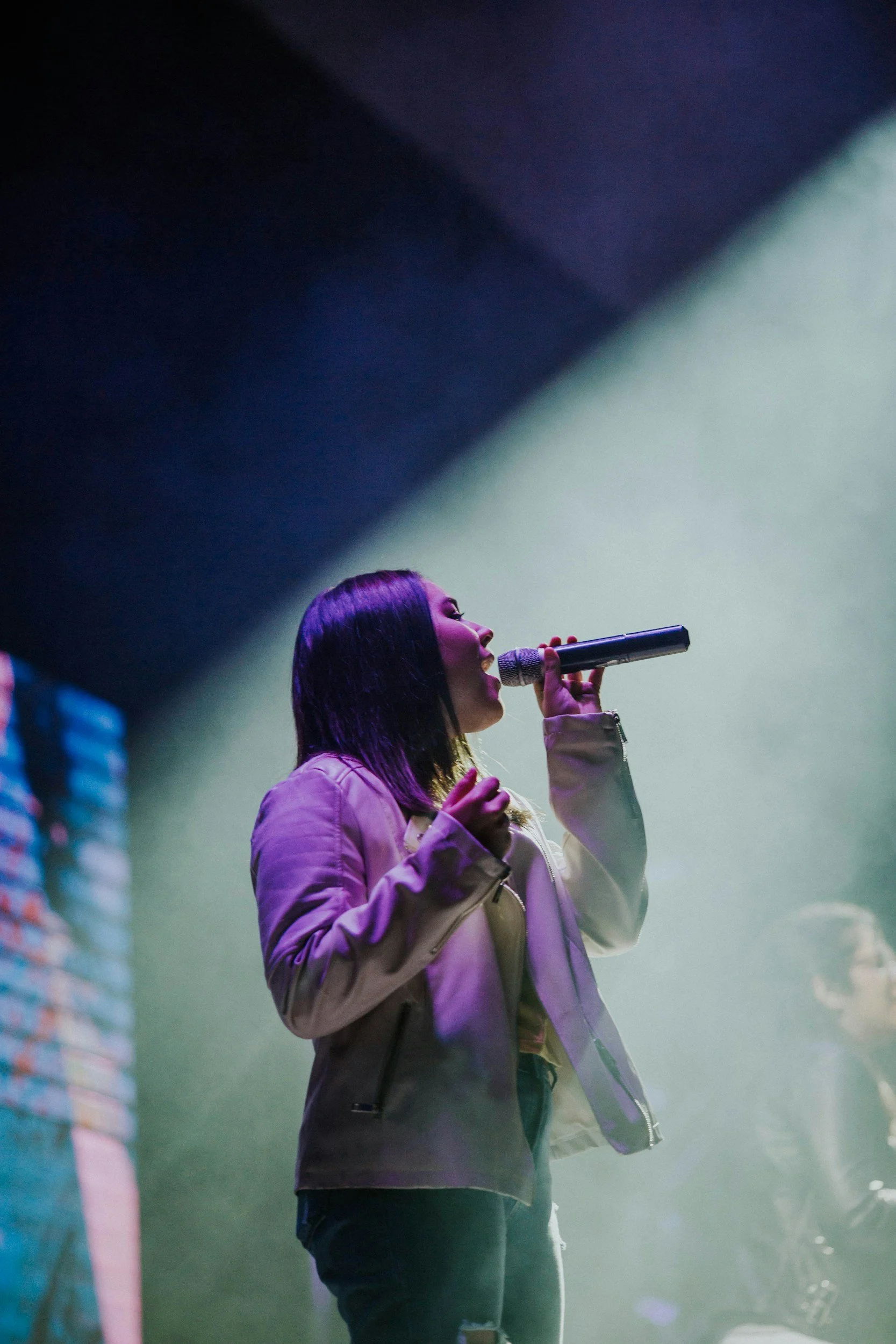 A woman with dark purple hair singing into a microphone on stage, illuminated by colorful stage lights.