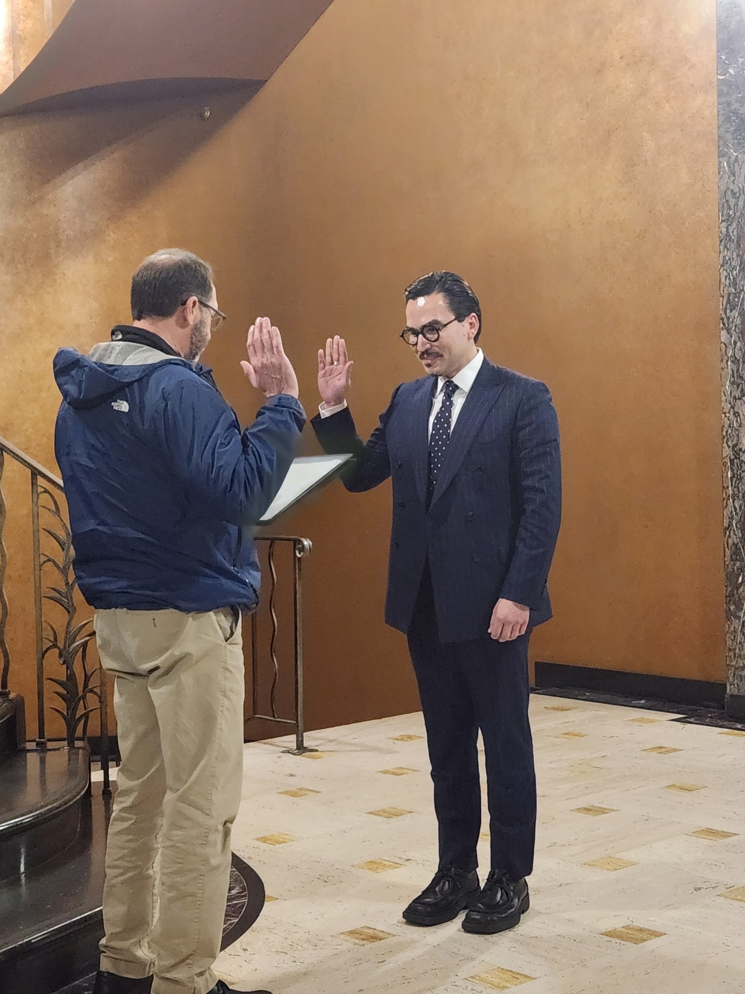 Two men are taking a professional oath, with one man in a suit and the other holding a clipboard, raising their right hands for a high five in an indoor setting.