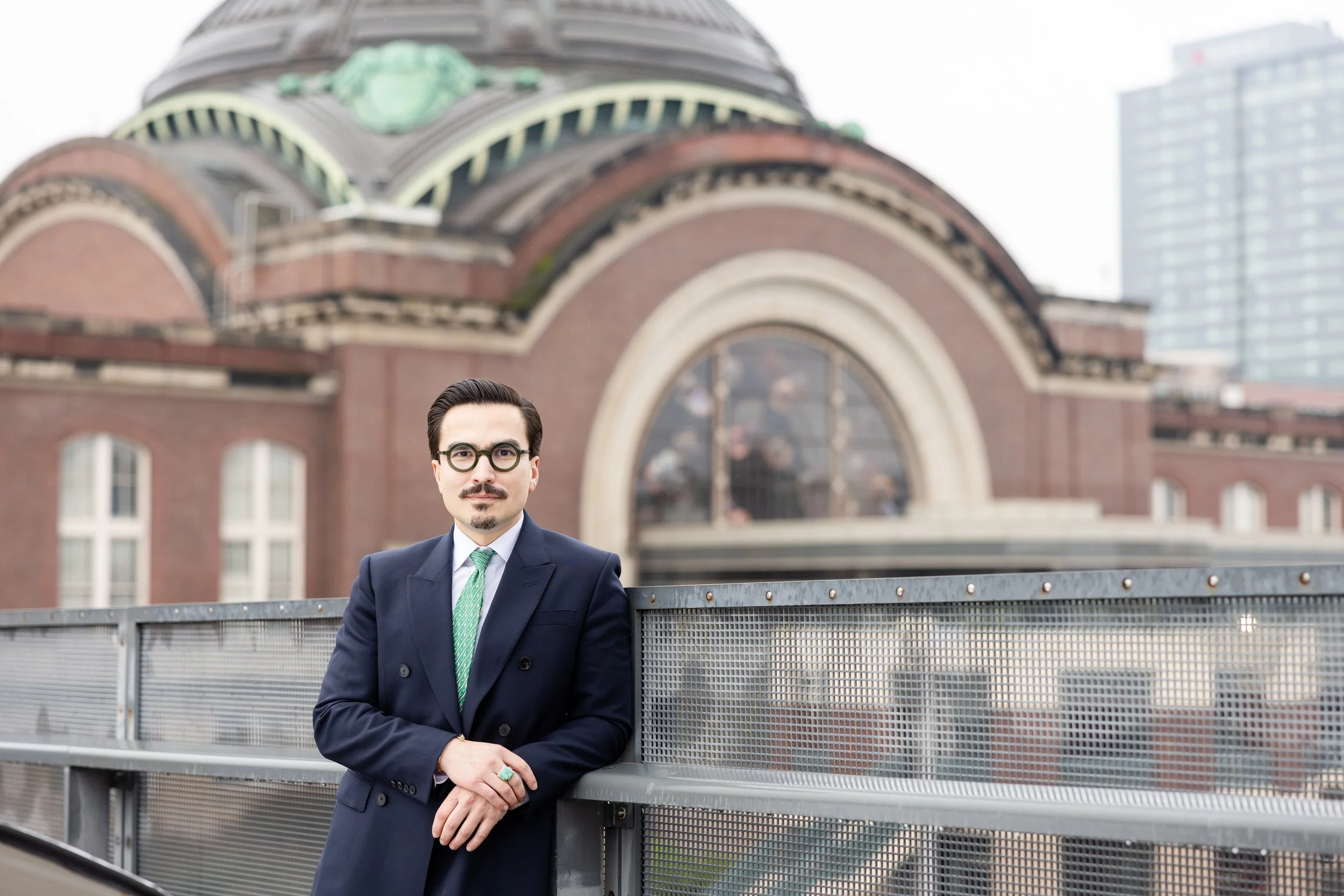 Sergio Flores is  dressed in a suit and tie standing outdoors on a city rooftop with a historic building and modern skyscrapers in the background.