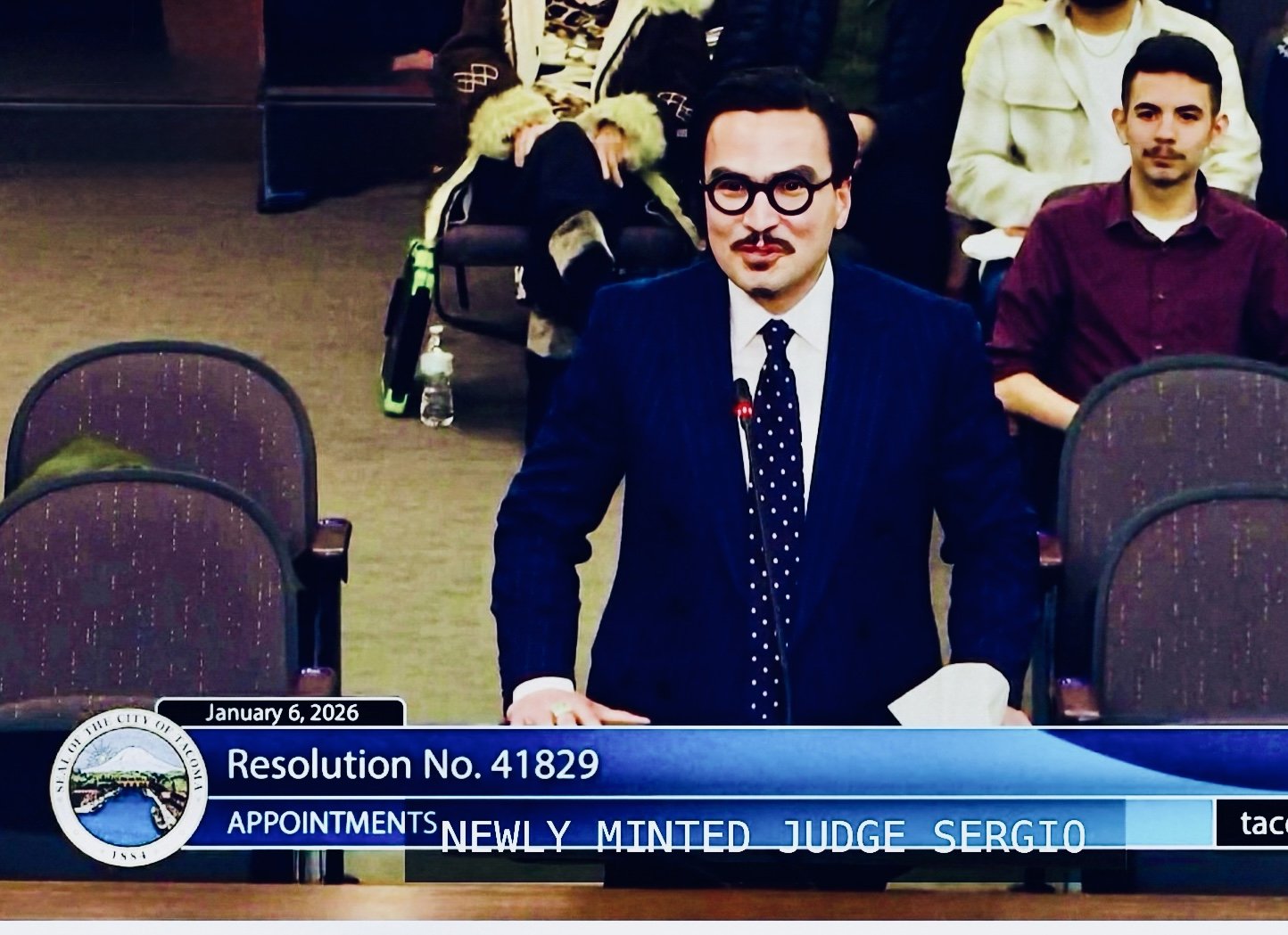 A man in a suit and glasses standing behind a desk with a microphone, at a formal meeting or hearing. Behind him are seated people, and a digital display shows a resolution and text indicating he is Judge Sergio. The digital overlay displays the date as January 6, 2026, and the resolution number 41829.