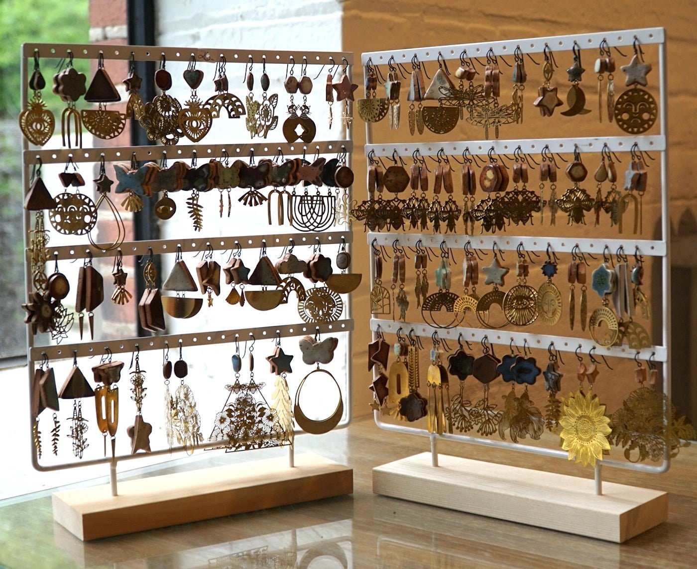 Two display racks filled with a variety of earrings, set on a wooden table near a window with a brick wall in the background.