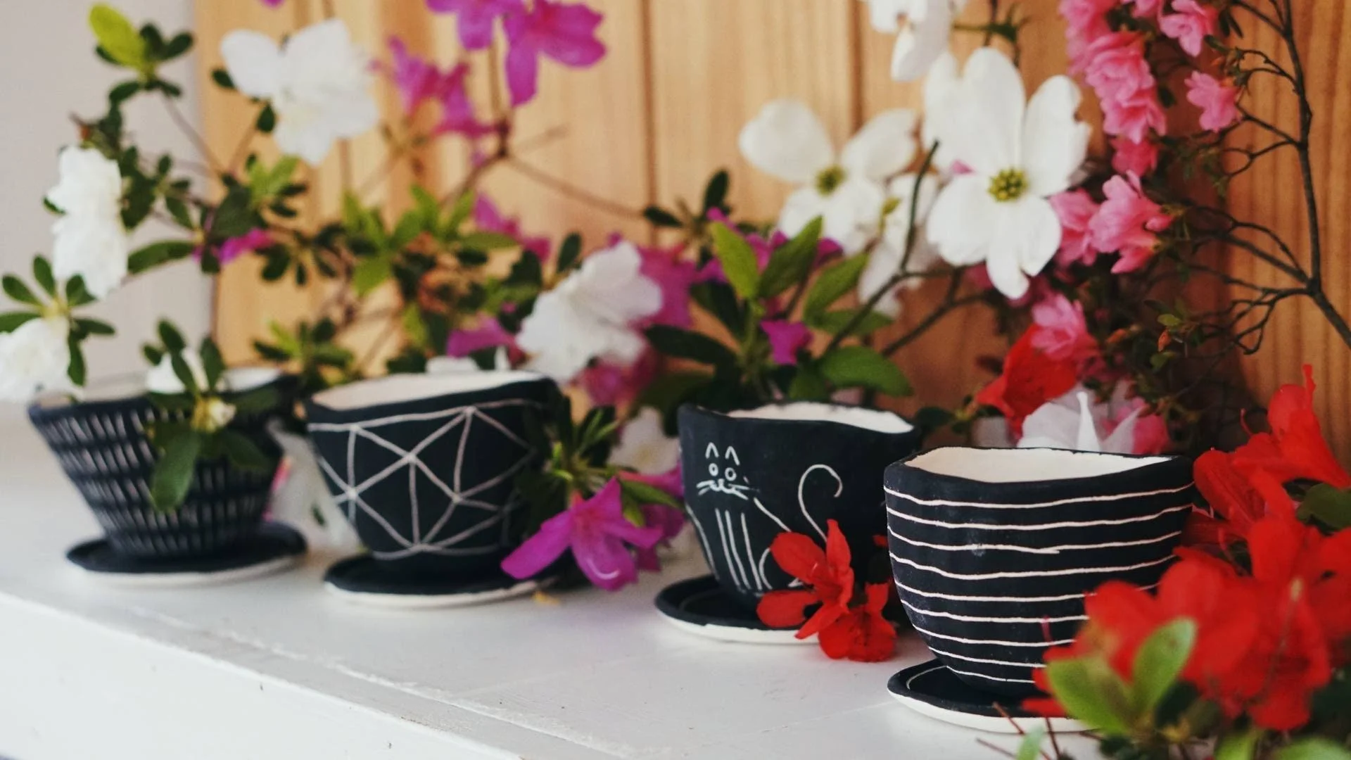 A row of four black and white ceramic cups with botanical and animal designs, placed on a white surface with colorful flowers in the background.