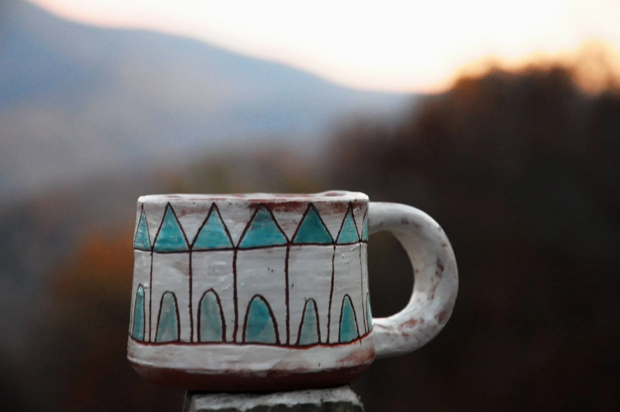 A ceramic mug with geometric patterns in blue and green on a white background, placed outdoors at sunset with blurred natural landscape in the background.