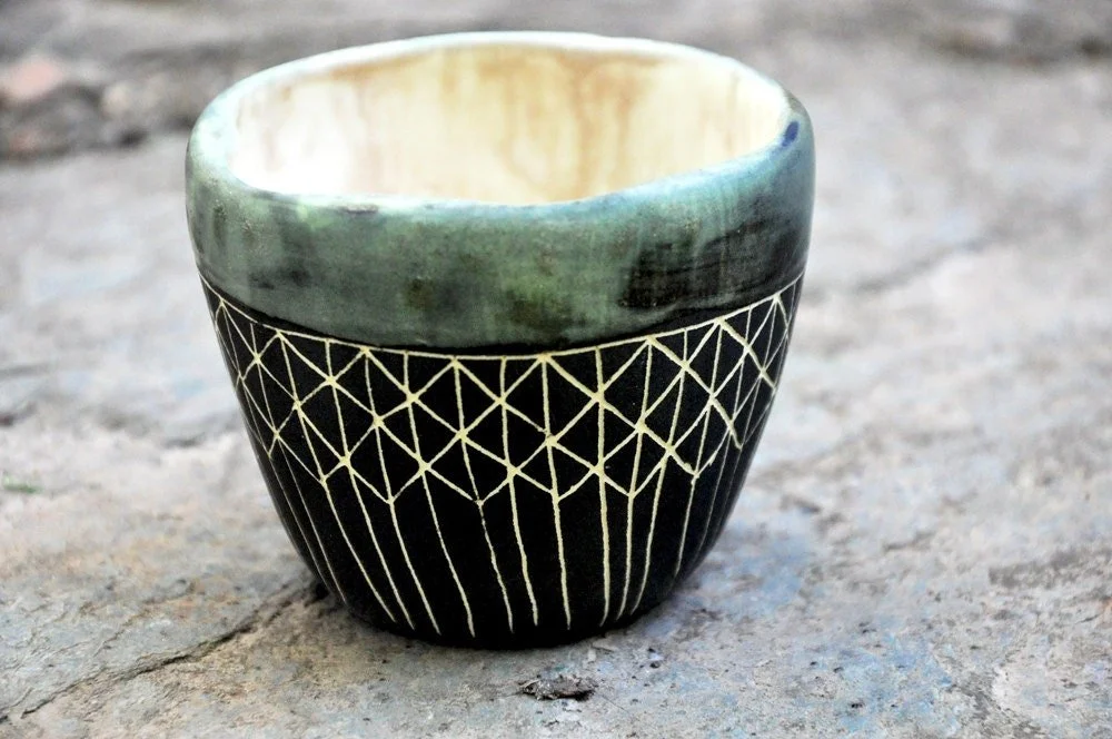 A small ceramic cup with intricate white geometric patterns on a black background, placed on a textured stone surface.