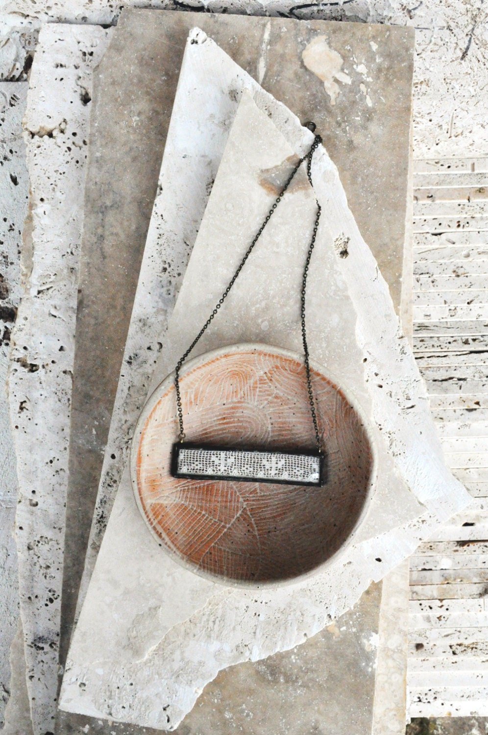 A necklace with a rectangular pendant hanging over a round, carved stone bowl placed on a flat stone surface.