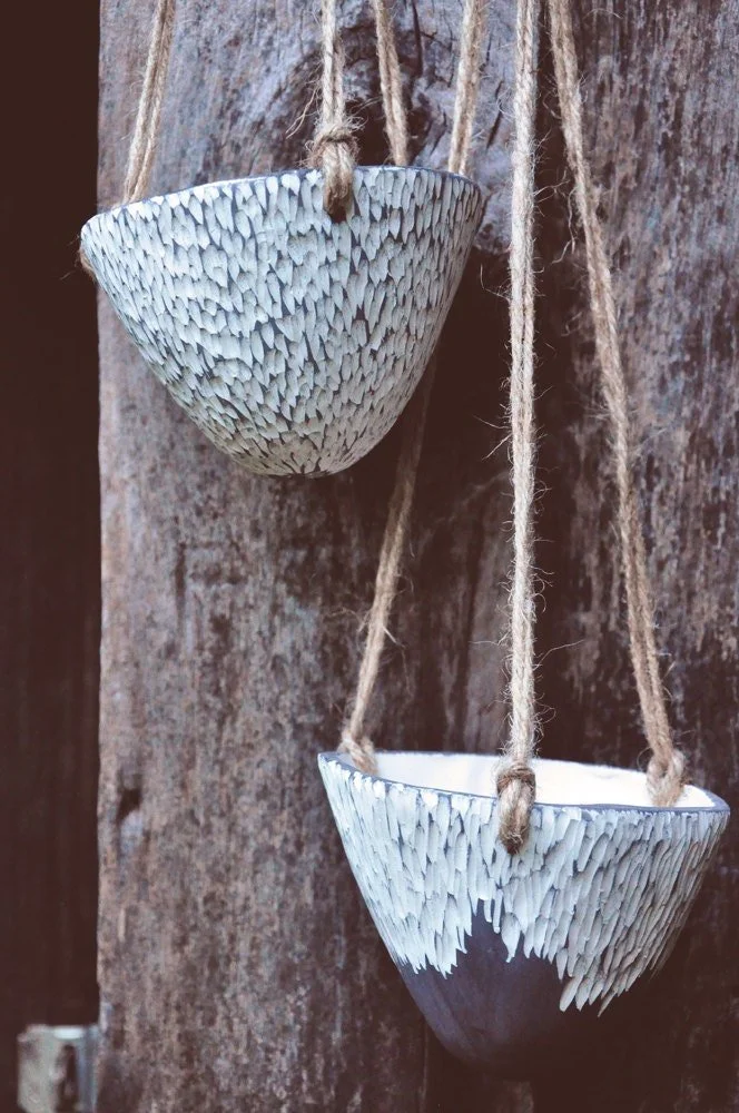 Two hanging ceramic bowls with textured white exterior, suspended by twine against a wooden background.