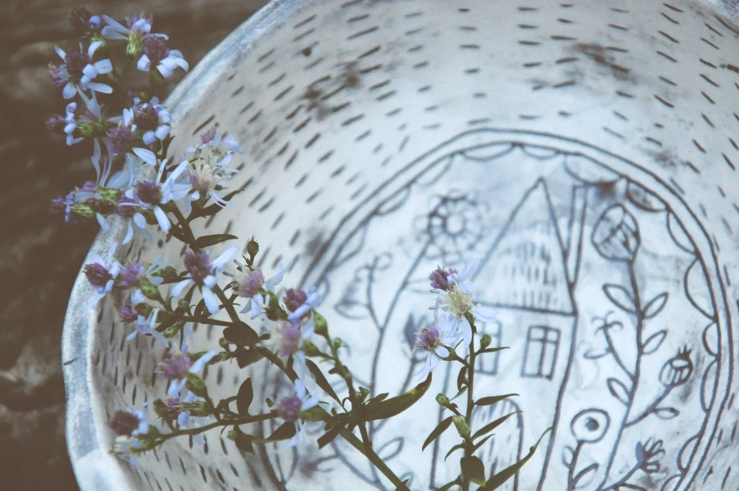 A decorative metal tray with a house and floral design, holding small purple and white flowers.