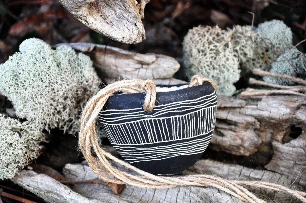 A black pottery bowl with white horizontal and vertical line patterns, decorated with a jute rope, surrounded by dried moss and wood on the ground.
