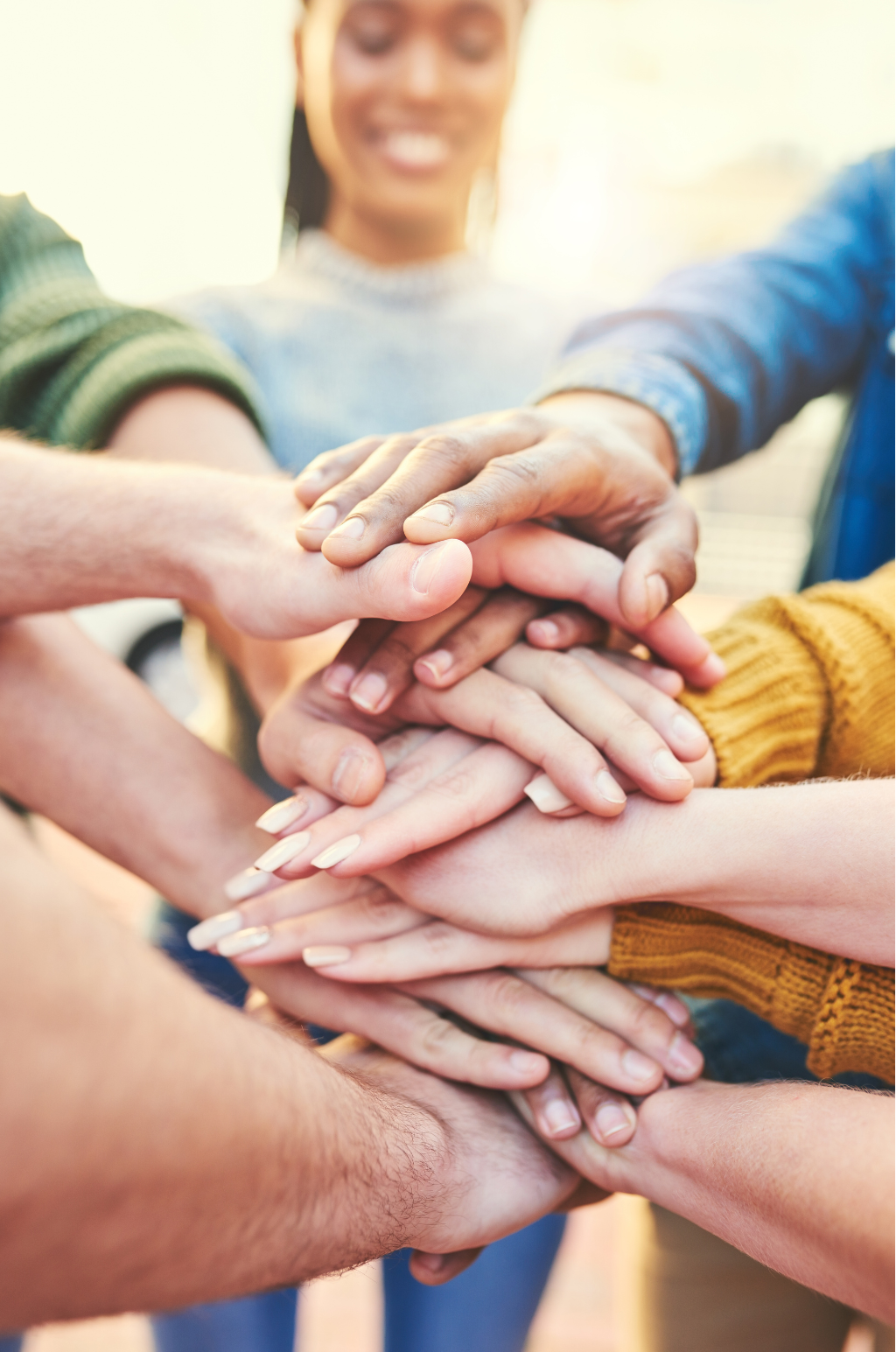 Multiple hands stacked together in teamwork gesture, diverse group of people, close-up, casual clothing, smiling woman in background.
