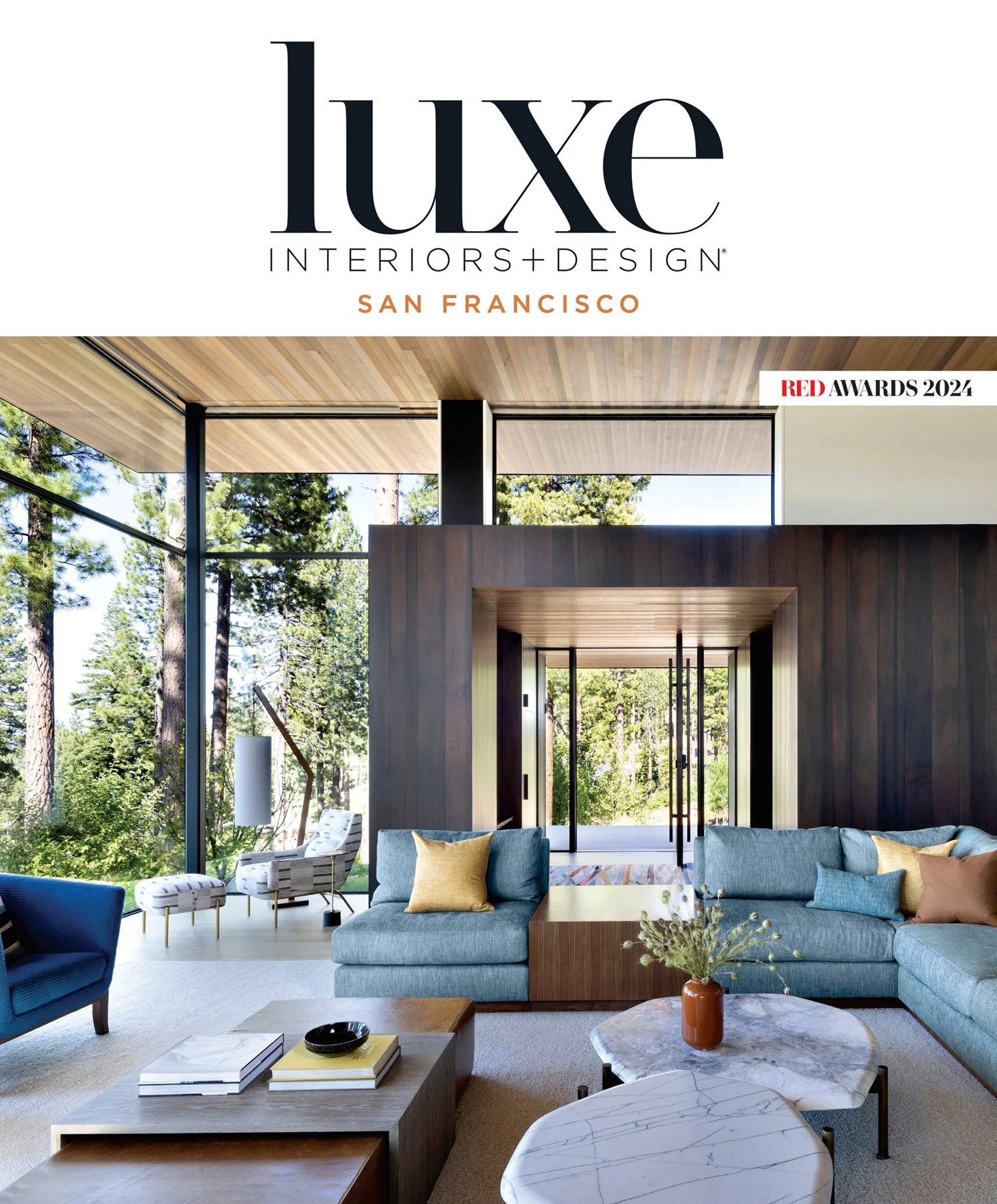 Interior of a modern living room with large windows, blue sofas, a marble coffee table, and wooden accents, overlooking a forest.