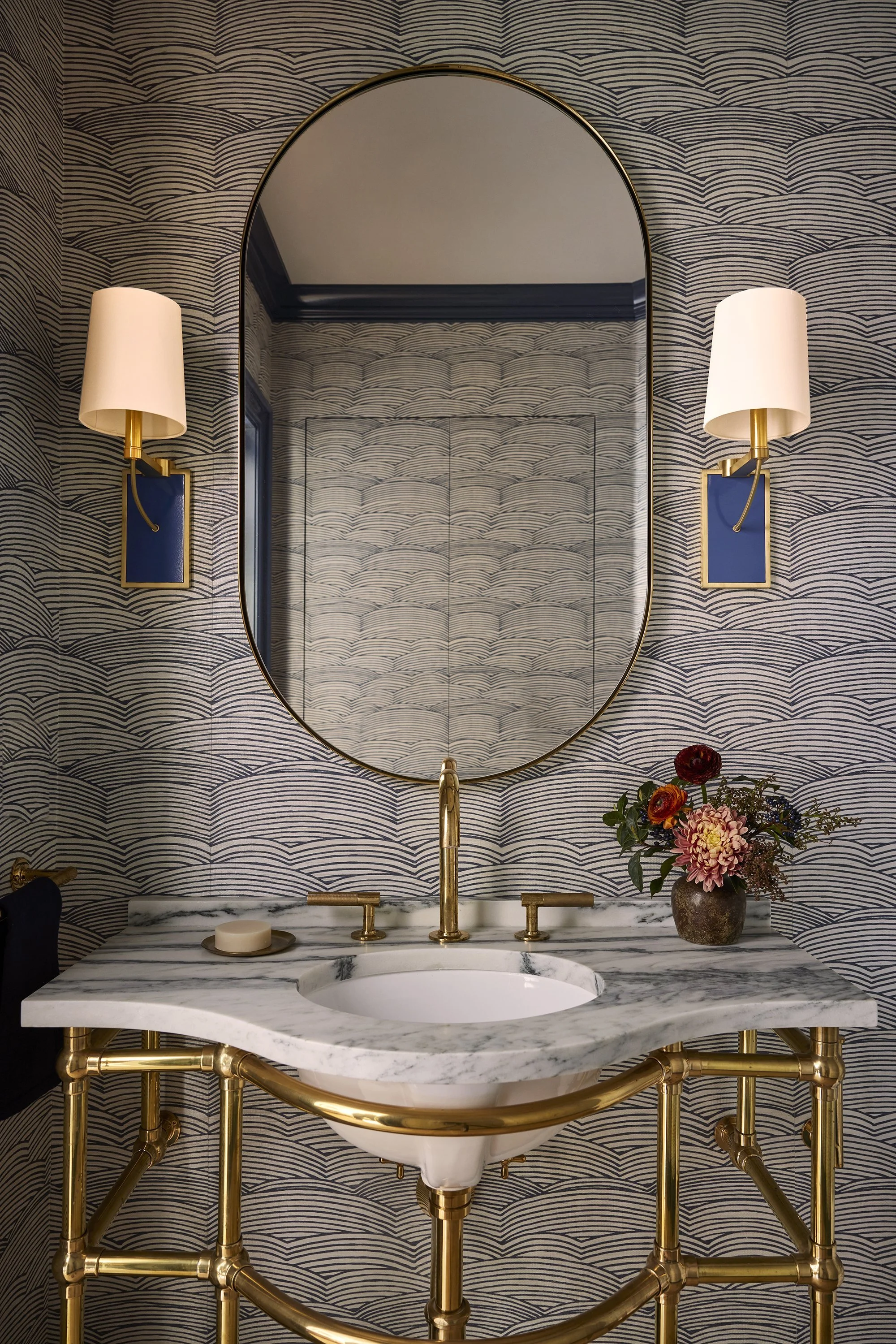 An elegant bathroom vanity with a marble countertop, gold fixtures, a large oval mirror, two wall sconces with white shades, a vase of flowers, and patterned wallpaper.