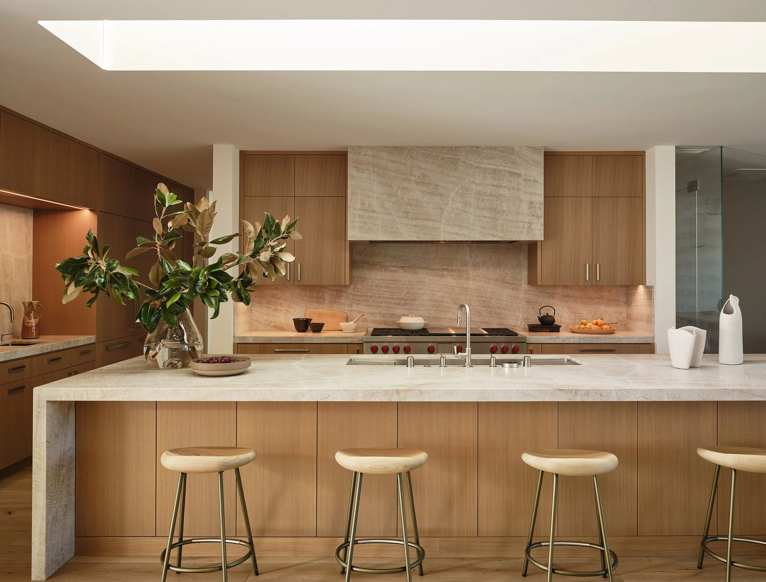 Modern kitchen with beige marble island, wooden cabinetry, and a skylight. Decor includes a glass vase with green leaves, a bowl of plums, white and black bowls, a teapot, a plate of oranges, and white vases. Wooden barstools line the island.