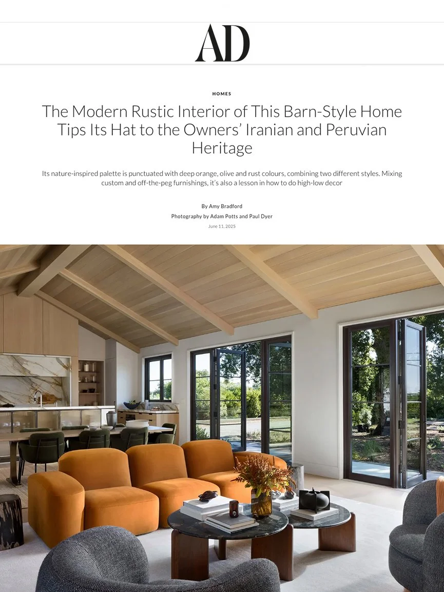 Cover page of a magazine or article titled "The Modern Rustic Interior of This Barn-Style Home Tips Its Hat to the Owners' Iranian and Peruvian Heritage" Showing a spacious living room with large windows, orange and gray chairs, a black round coffee 