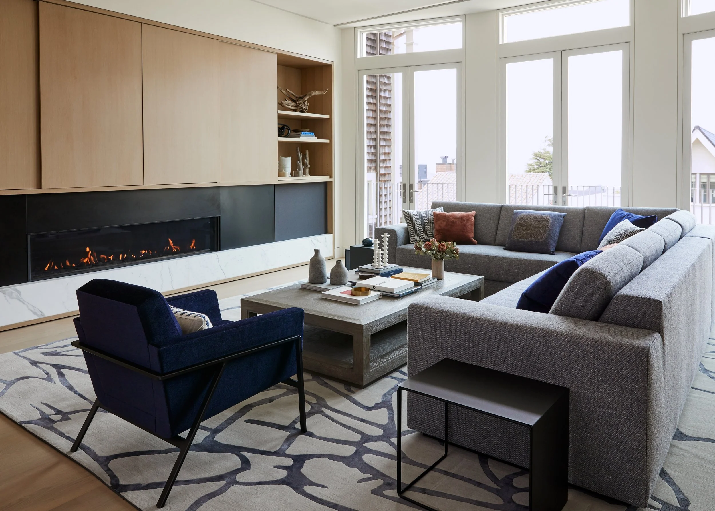 Modern living room with large windows, gray sectional sofa with colorful pillows, black armchair, table with decorative items, and a built-in fireplace with a marble surround.
