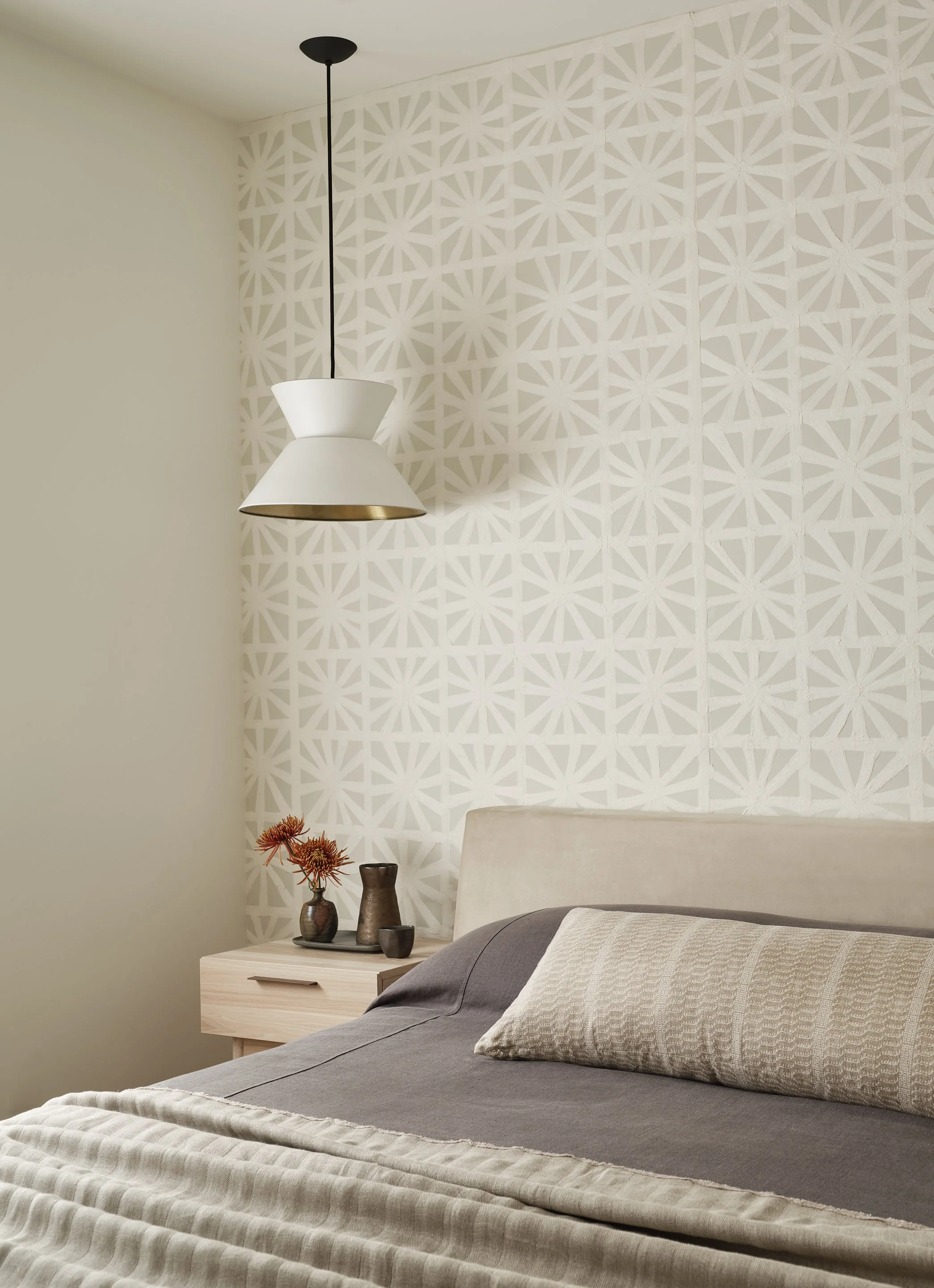 A bedroom with a modern white pendant light, a textured beige headboard, a gray bedspread, a decorative pillow, and a nightstand with vases and flowers. The wall behind the bed features a geometric patterned wallpaper.