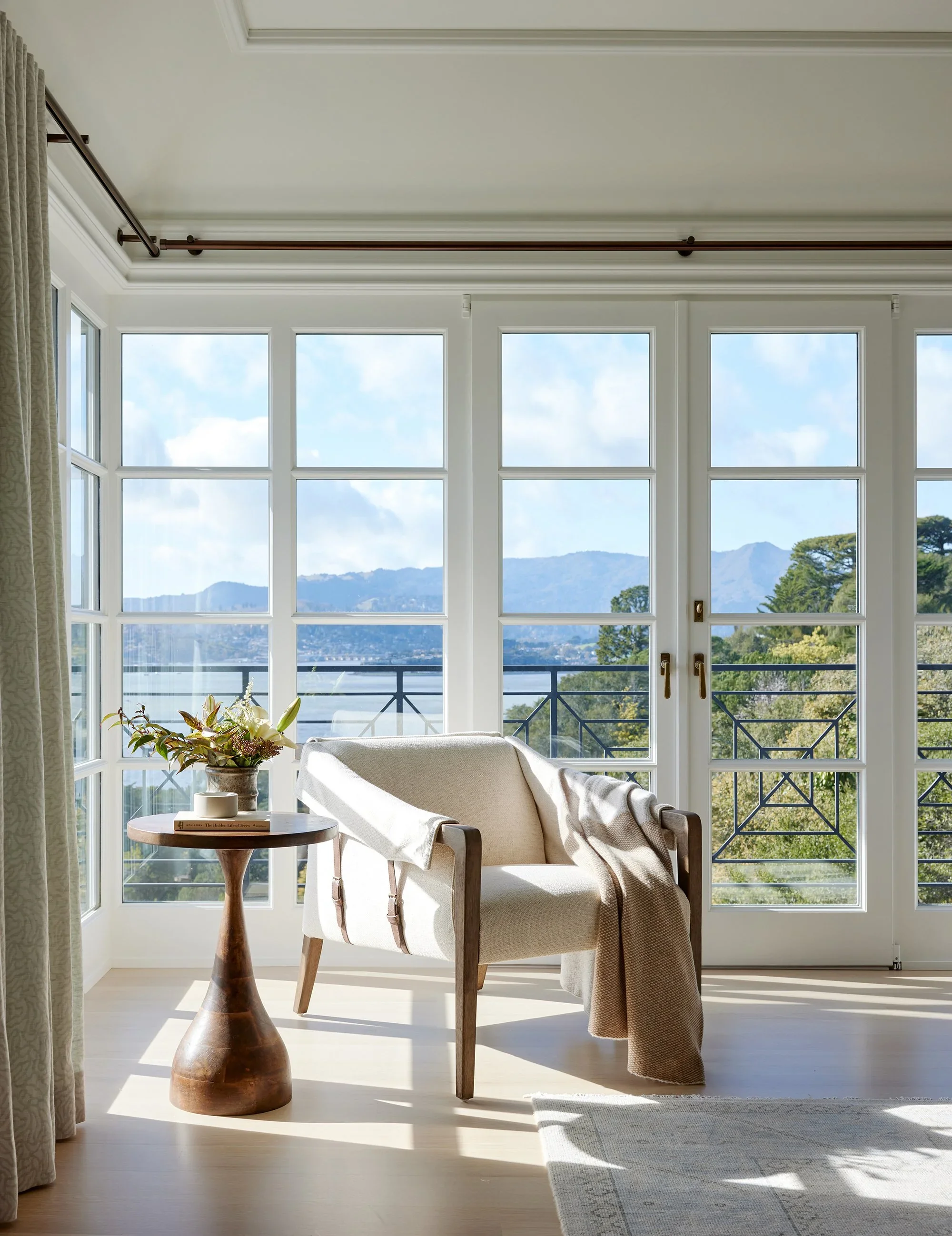 Sunlit room with large windows and balcony view of mountains, furnished with a beige armchair, a wooden side table with a plant, a book, and a small decorative object, and a throw blanket on the chair.