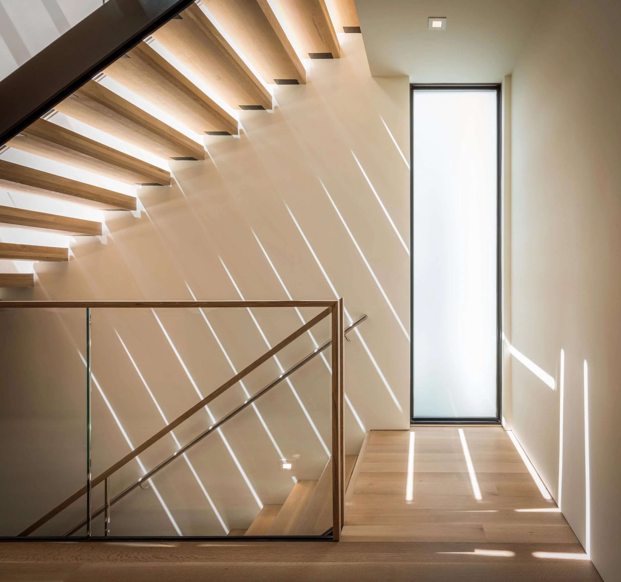 Sunlight streaming through a tall, narrow window creates diagonal light and shadow patterns on a white wall and wooden floor in a modern stairwell.