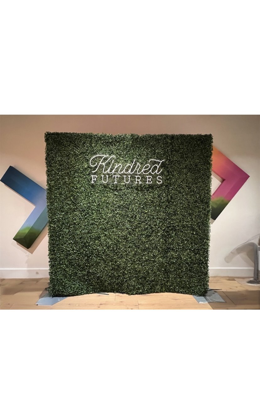 A decorative green hedge wall with the words "Kindred Futures" written in cursive and uppercase lettering.