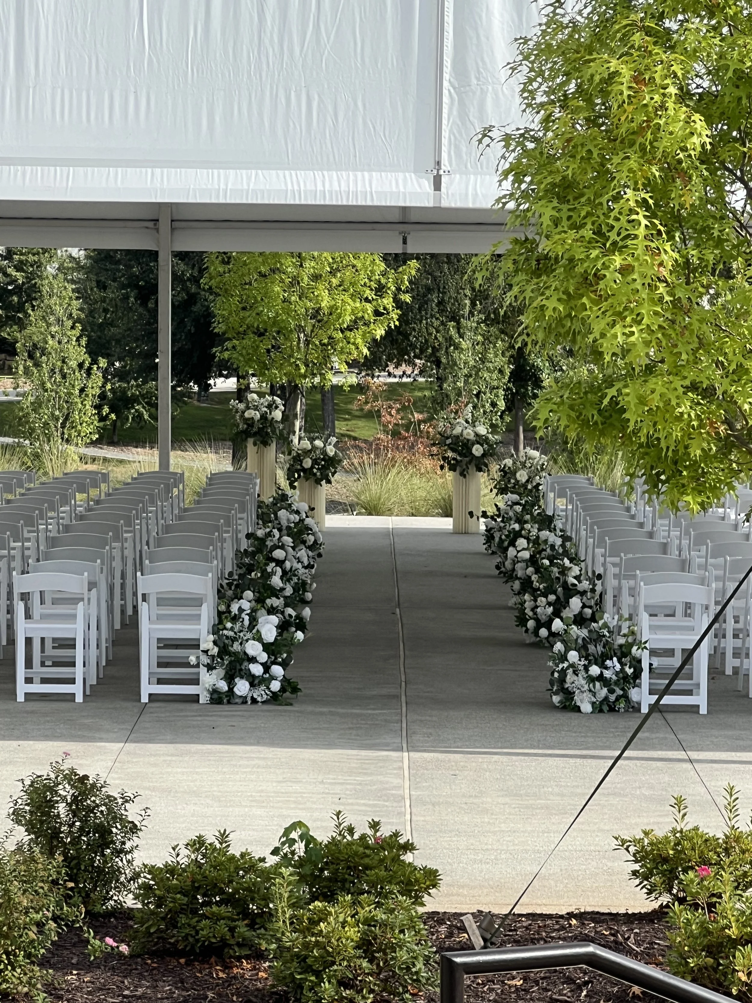 Outdoor wedding ceremony setup with white chairs arranged on each side of a flower-decorated aisle, under a white canopy, surrounded by green trees and shrubs.