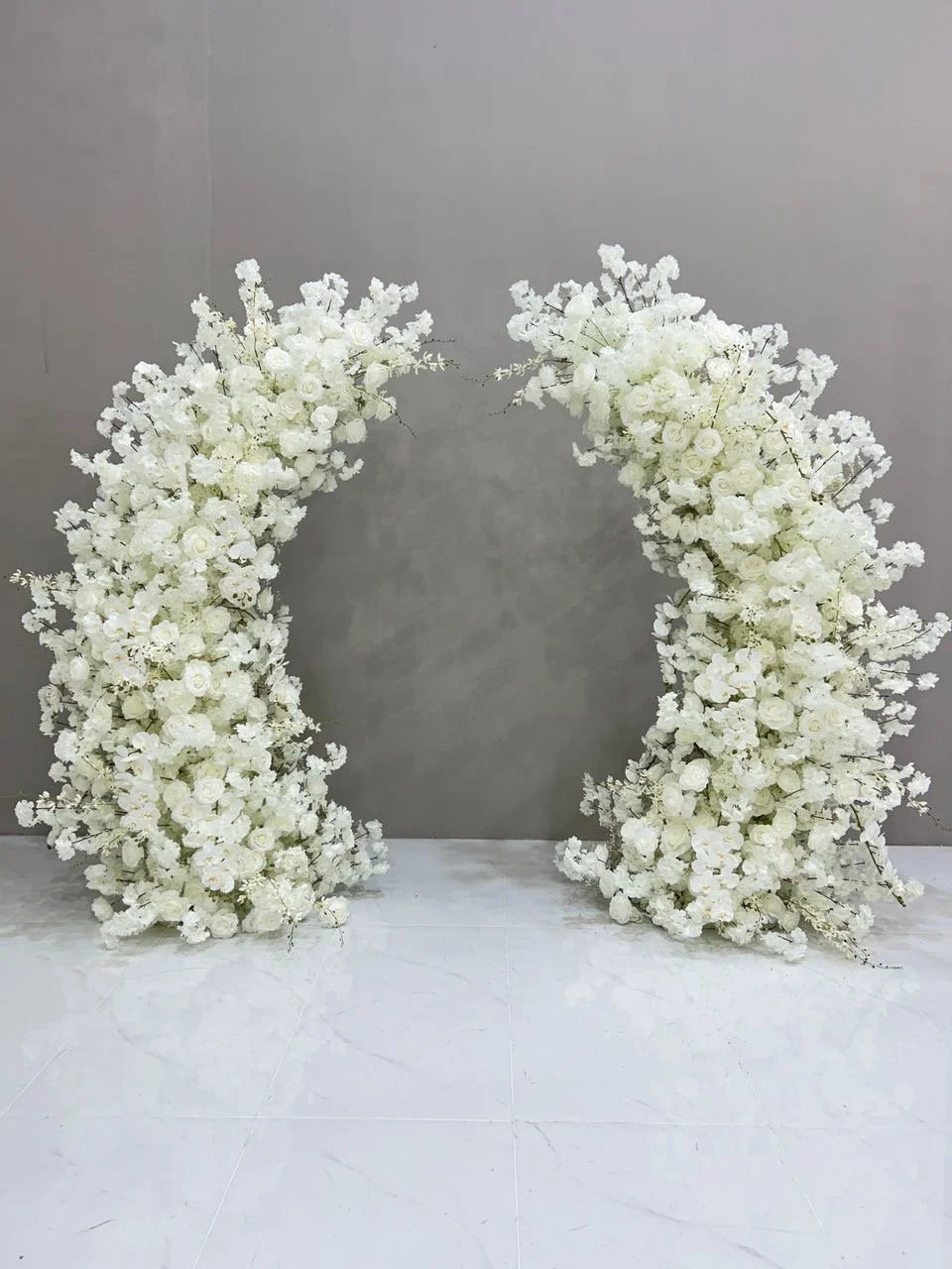 Large white floral arch made of white roses and other flowers, set against a gray wall for a special event or wedding.