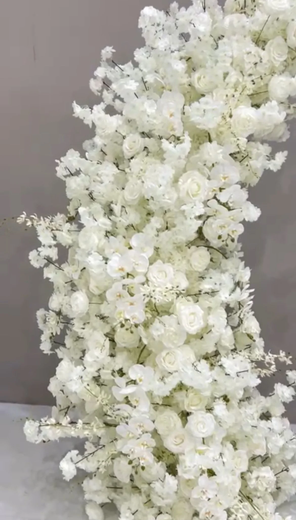 A large arrangement of white flowers, including roses and orchids, displayed vertically against a gray background.