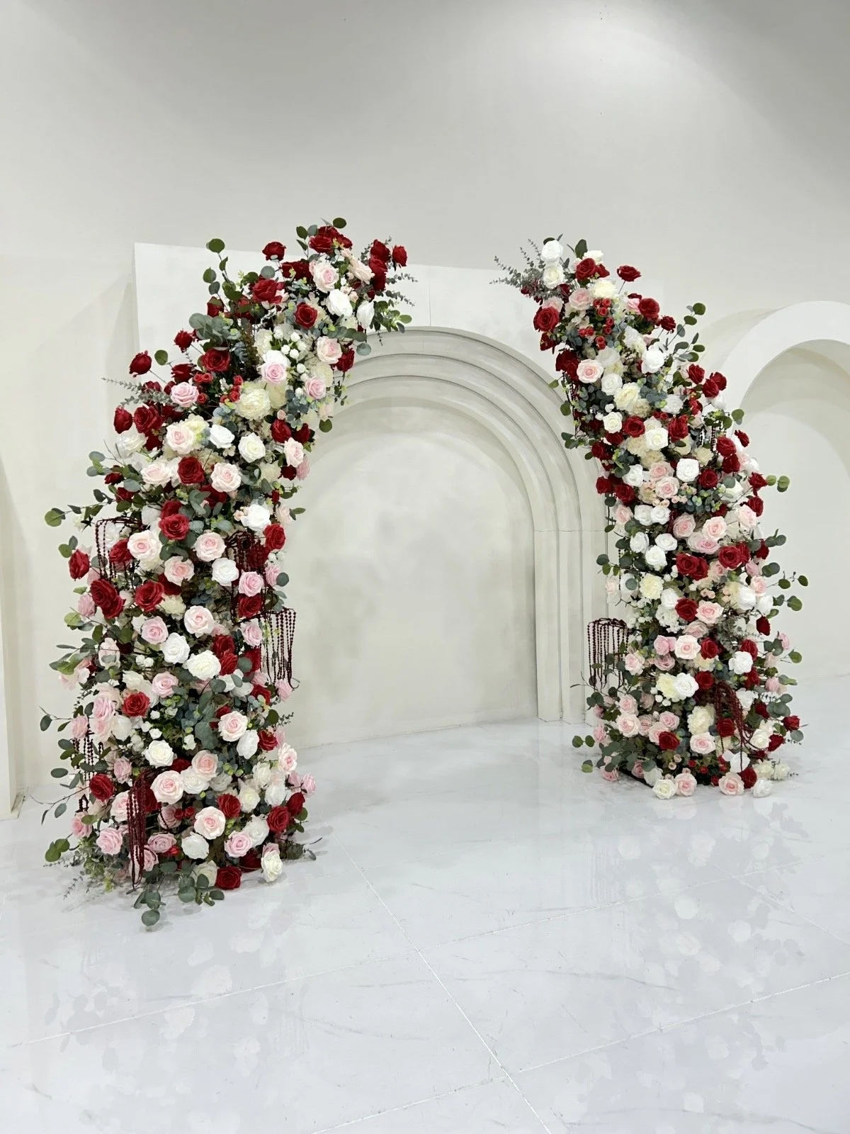 Flower arch with red, pink, and white roses and greenery standing in front of a white wall and white marble floor, likely for a wedding or event setup.