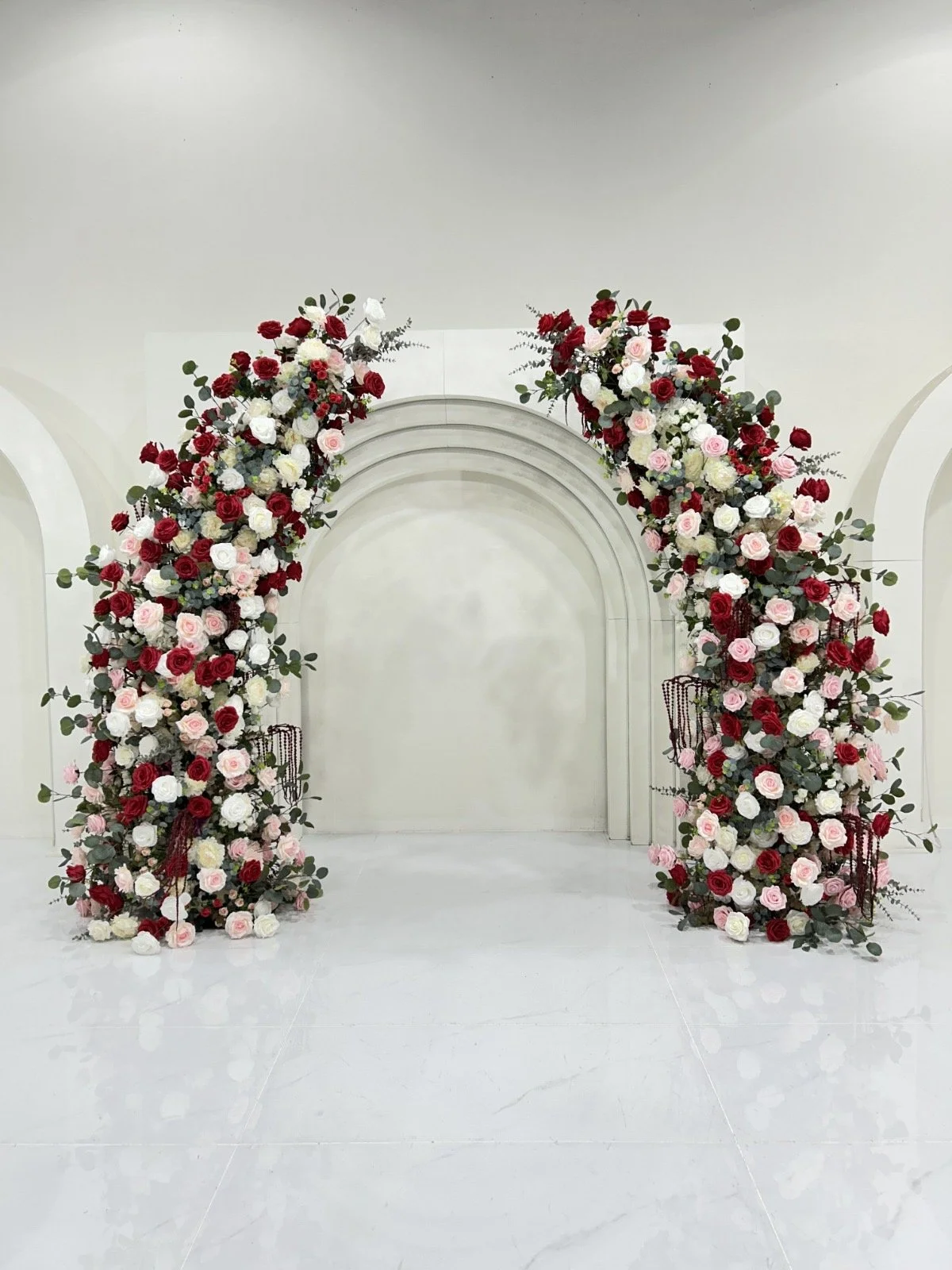 Floral arch with red, white, and pink roses and greenery on a white backdrop, used for wedding or event decoration.