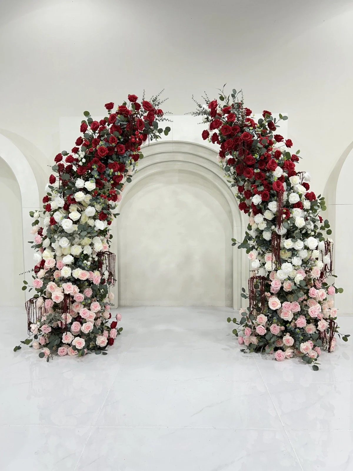 A floral wedding arch decorated with red, white, and pink roses, greenery, and hanging red beads, set against a minimalistic white backdrop.