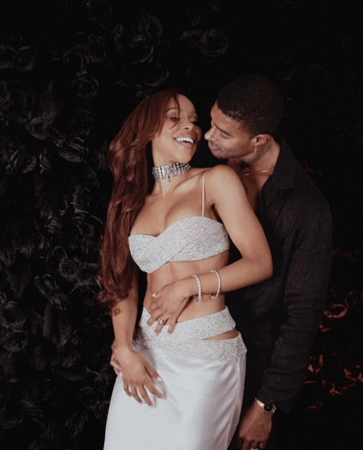 A woman in a sparkling silver evening outfit with a matching choker necklace and bracelet, smiling and leaning into a man in a black shirt, against a dark floral backdrop.