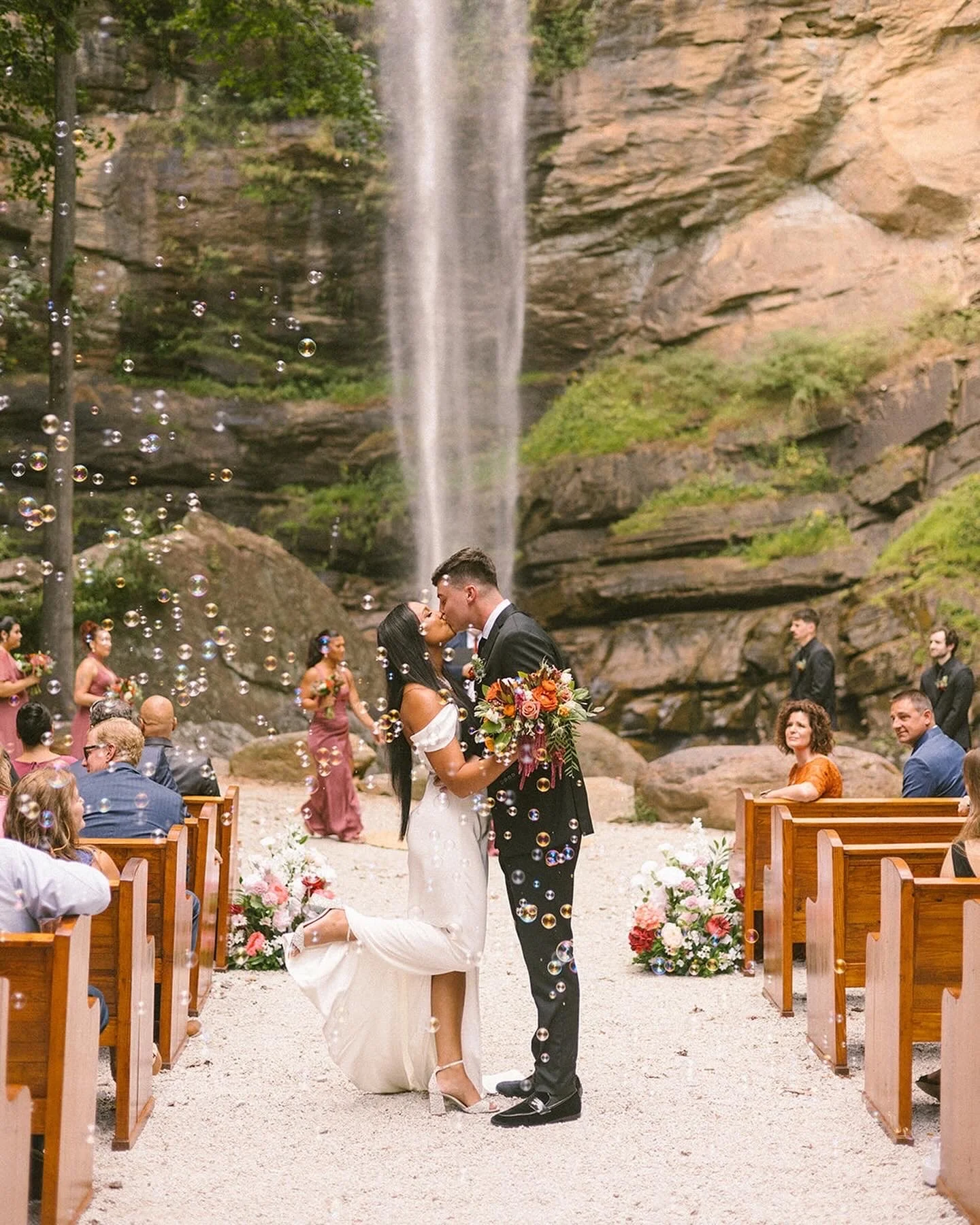 A couple kiss at their wedding ceremony outdoors under a waterfall, surrounded by guests and floral arrangements.