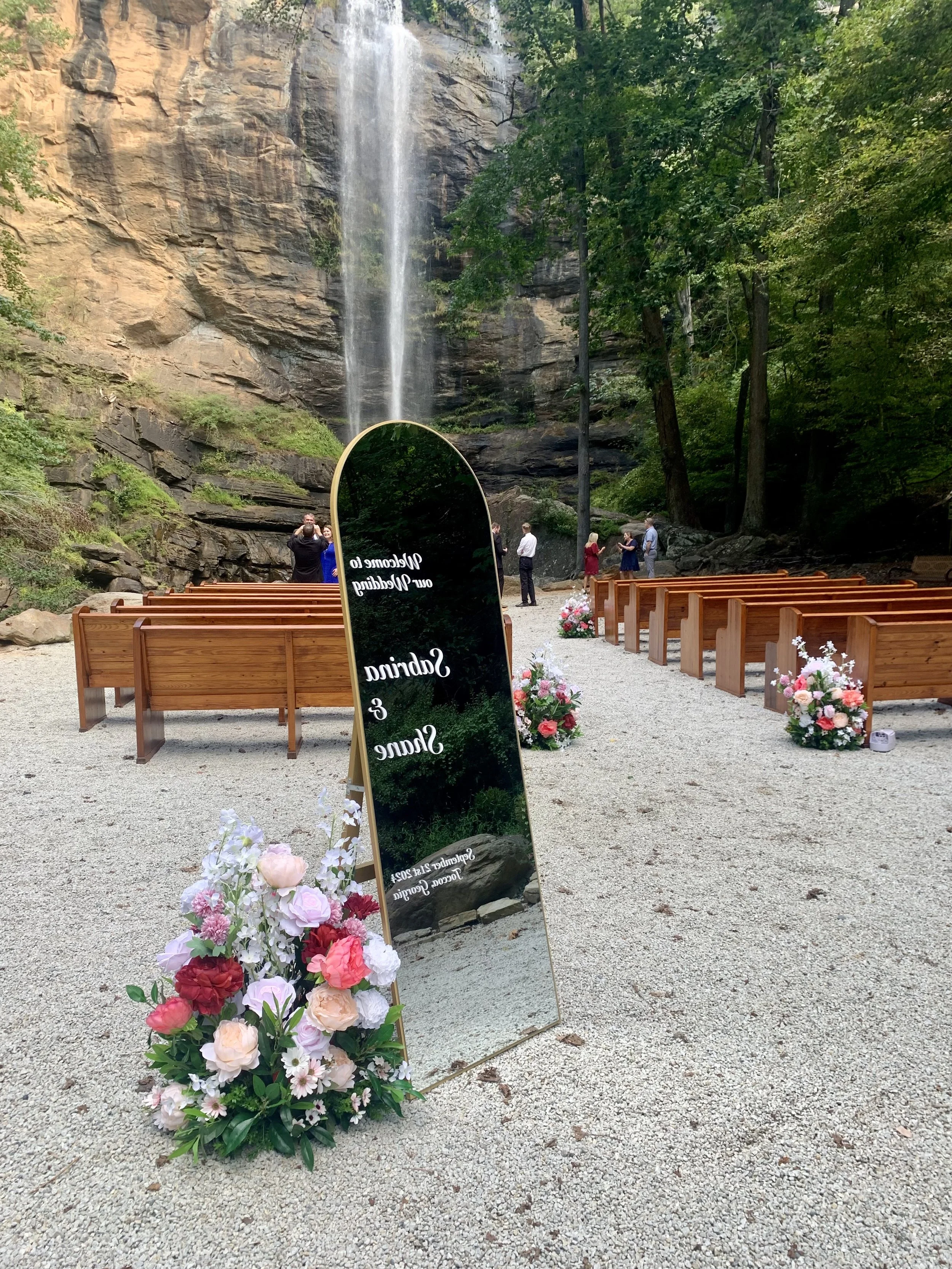 A wedding setup at a scenic outdoor location with a waterfall in the background, wooden benches, floral arrangements, and an angled mirror with wedding details reflected in it.