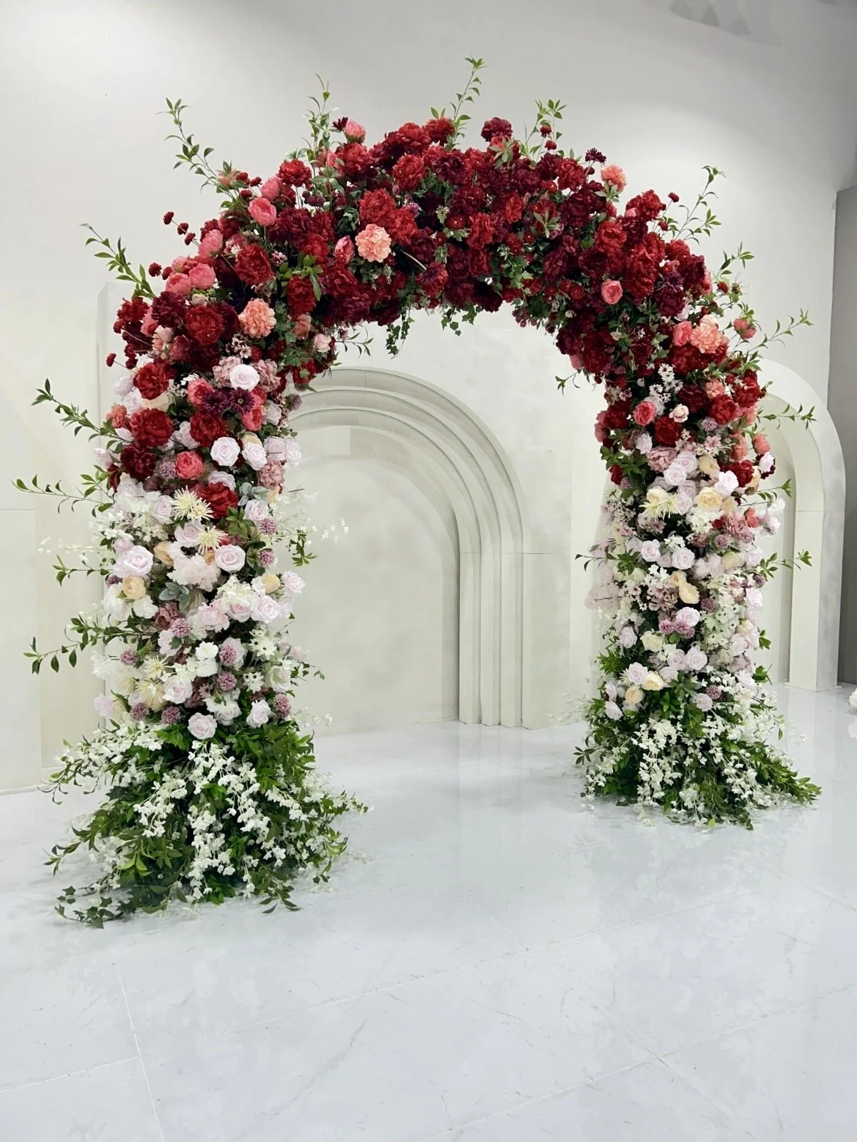 A floral arch made up of pink, red, and white roses and greenery, set in front of a white backdrop.