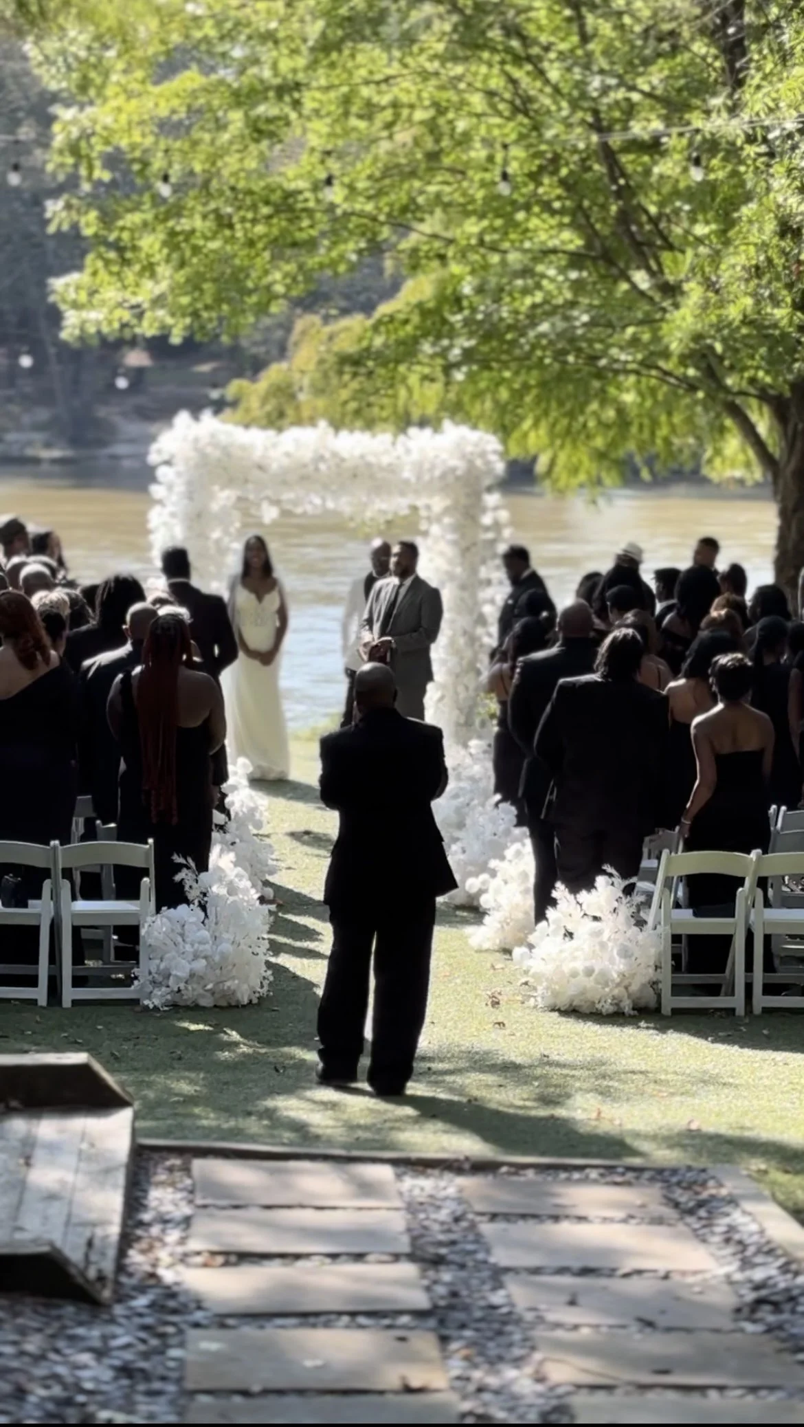 Wedding ceremony outdoors by a lake, with guests dressed in black, white, and gray, standing under a tree and facing the couple at the altar decorated with white flowers.