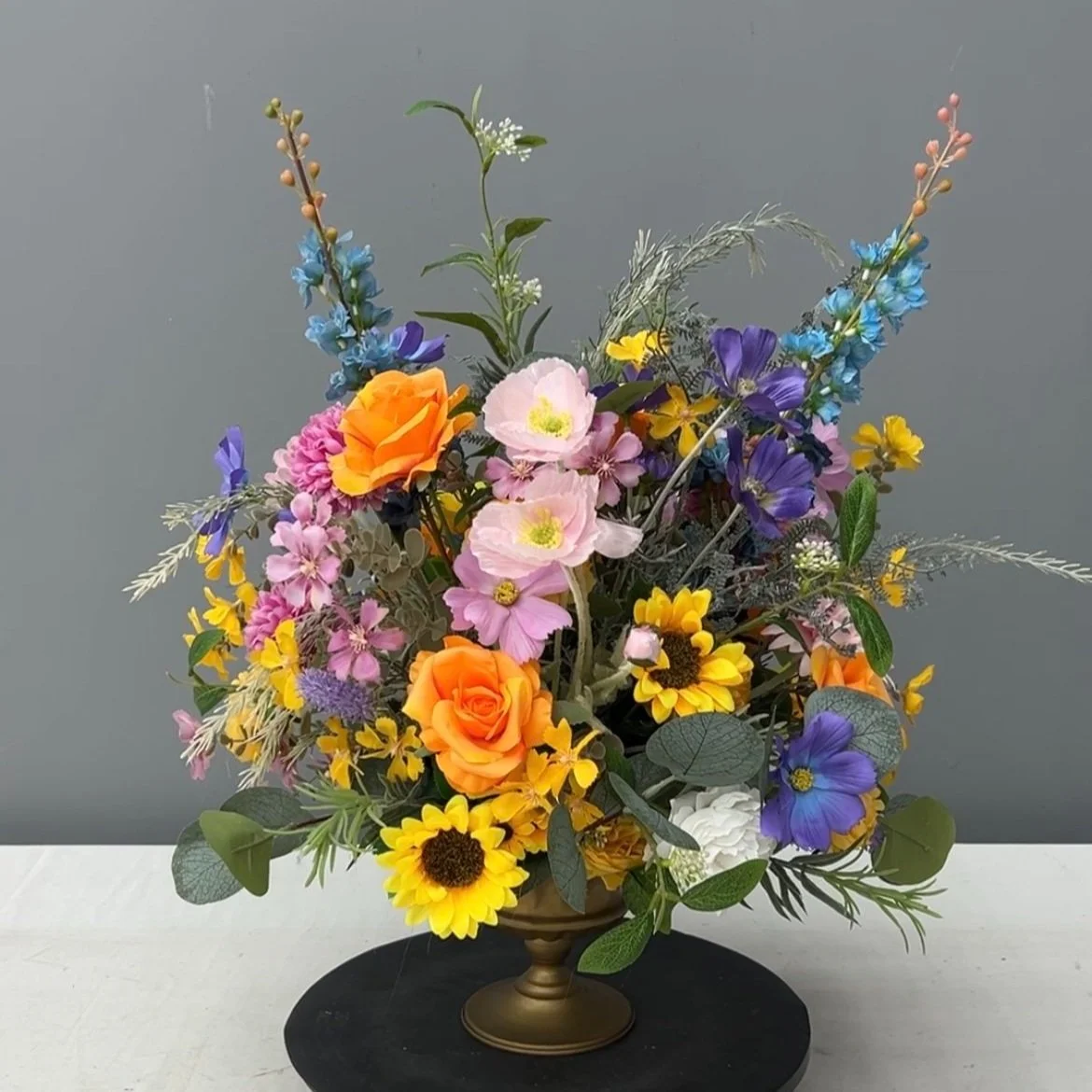Colorful flower bouquet in a gold vase on a white surface against a gray background.