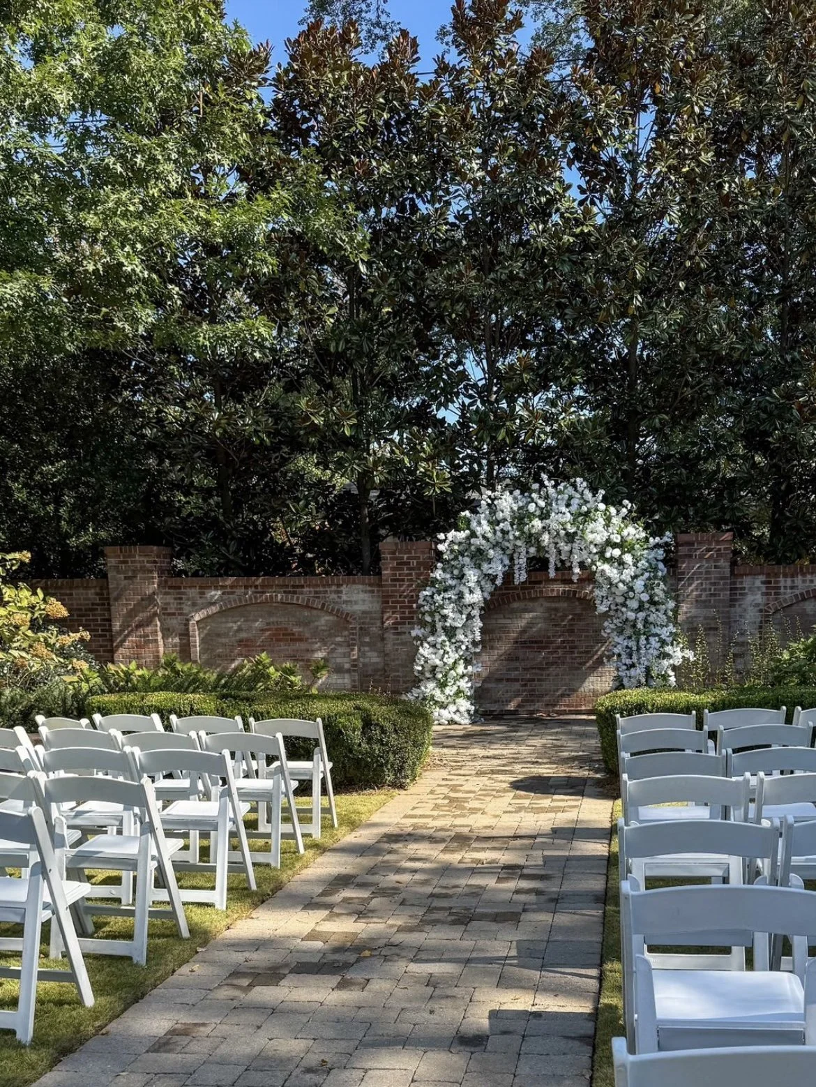 An outdoor wedding ceremony setup with white chairs arranged on either side of a brick pathway. At the end of the aisle, a floral arch made of white flowers is positioned against a brick wall, surrounded by lush greenery and trees under a clear blue sky.