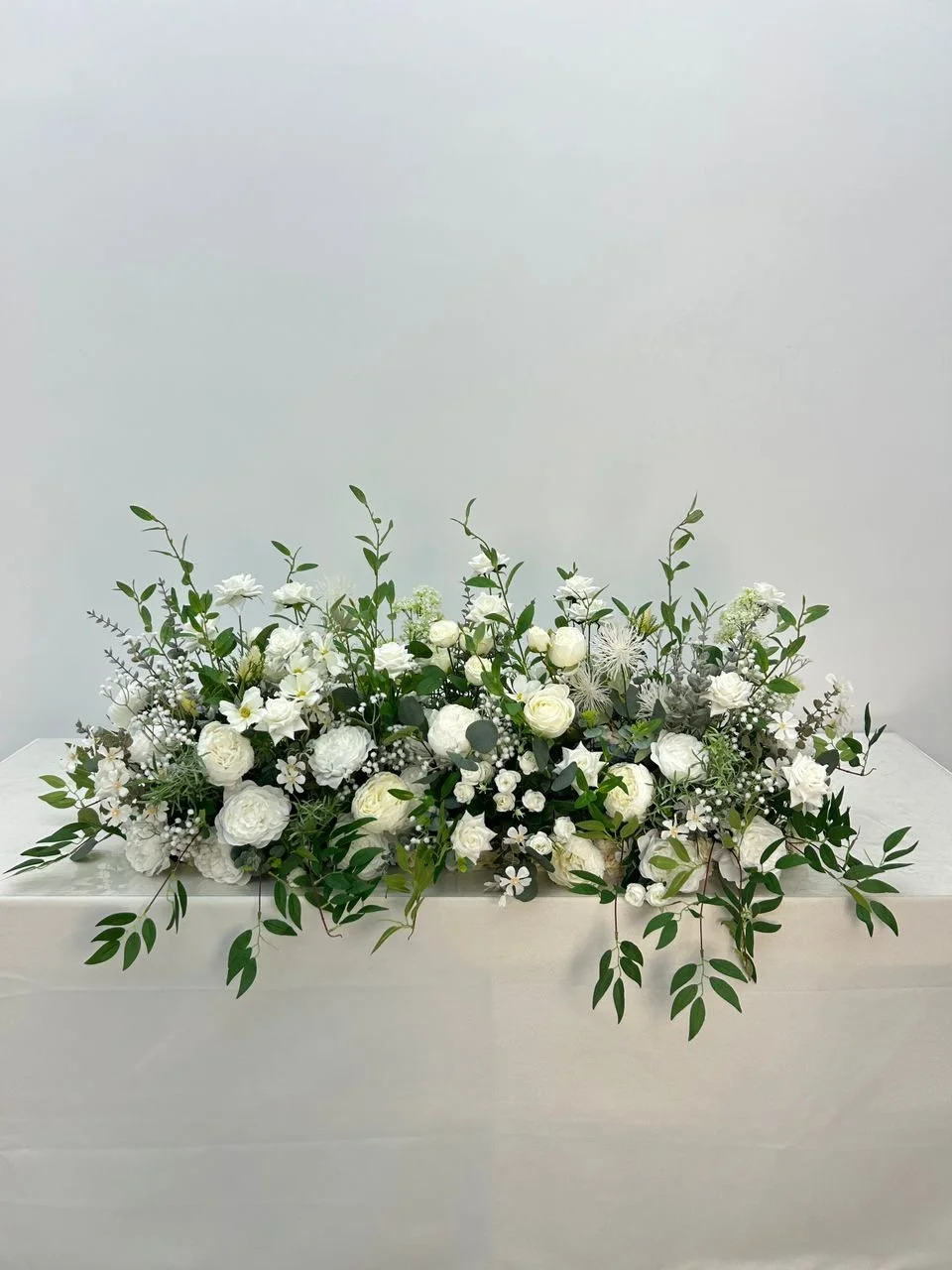 Arranged white floral display in a white container with various white flowers and greenery, set against a plain light-colored background.