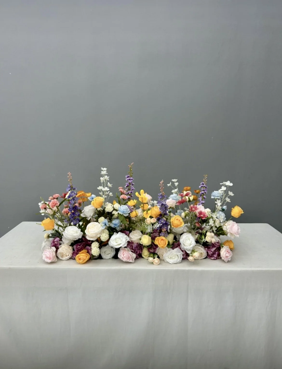 Colorful floral arrangement of roses, lavender, and other flowers on a white surface against a gray background.