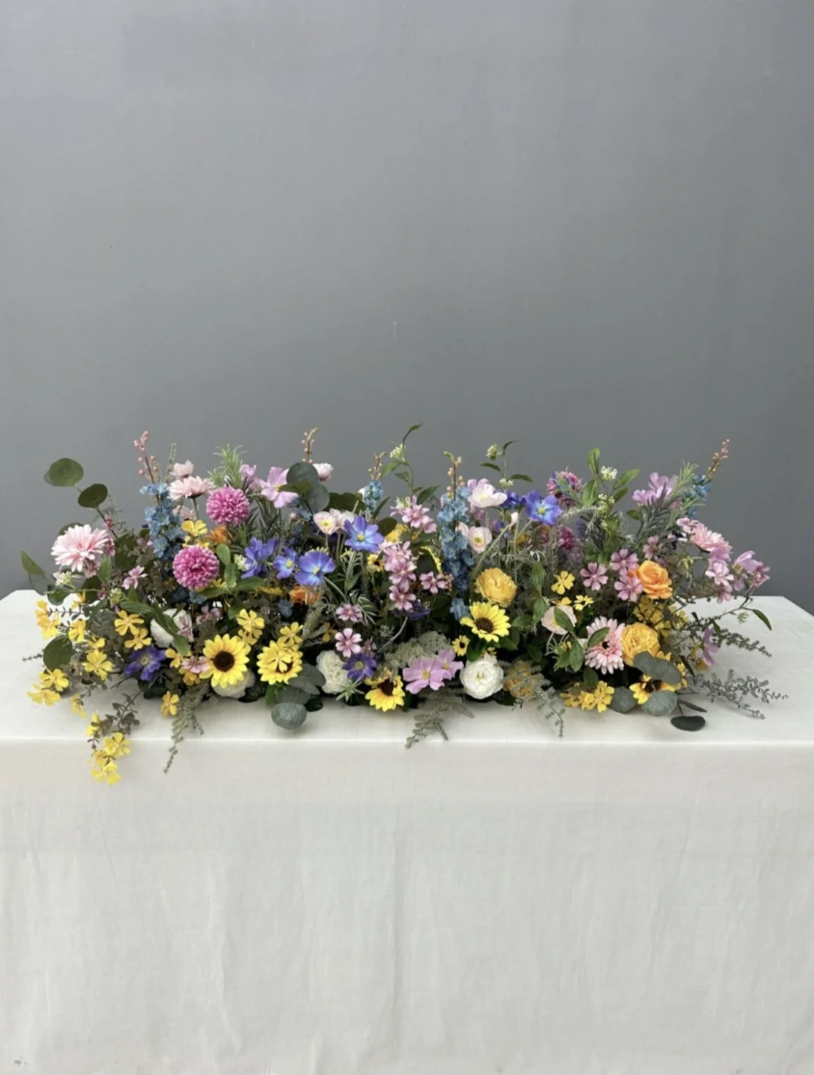 Colorful flower arrangement with pink, yellow, purple, and white flowers on a white table against a gray wall.