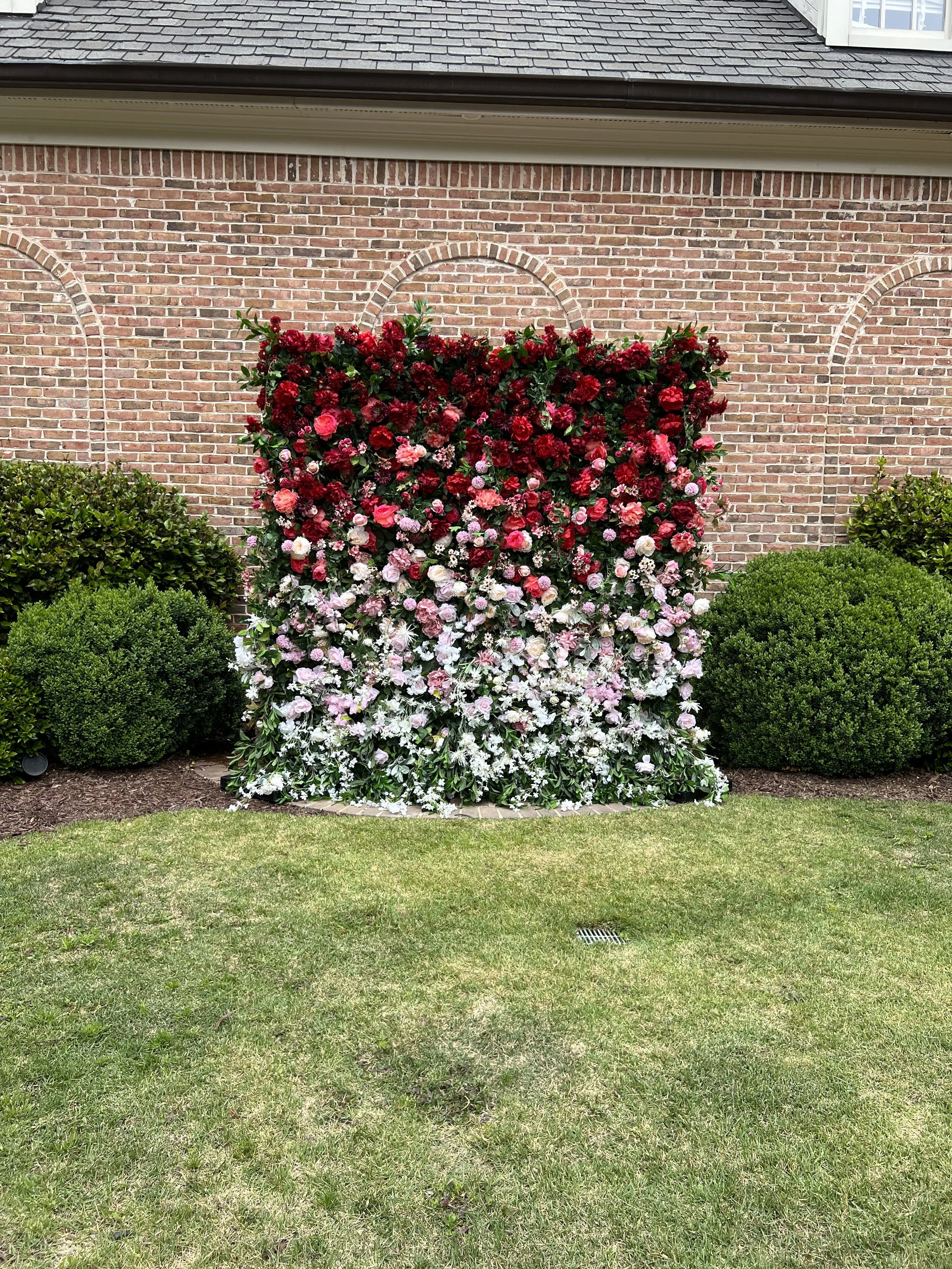 A decorative wall of red, pink, and white flowers in front of a brick house wall, with green shrubs and grass in the foreground.