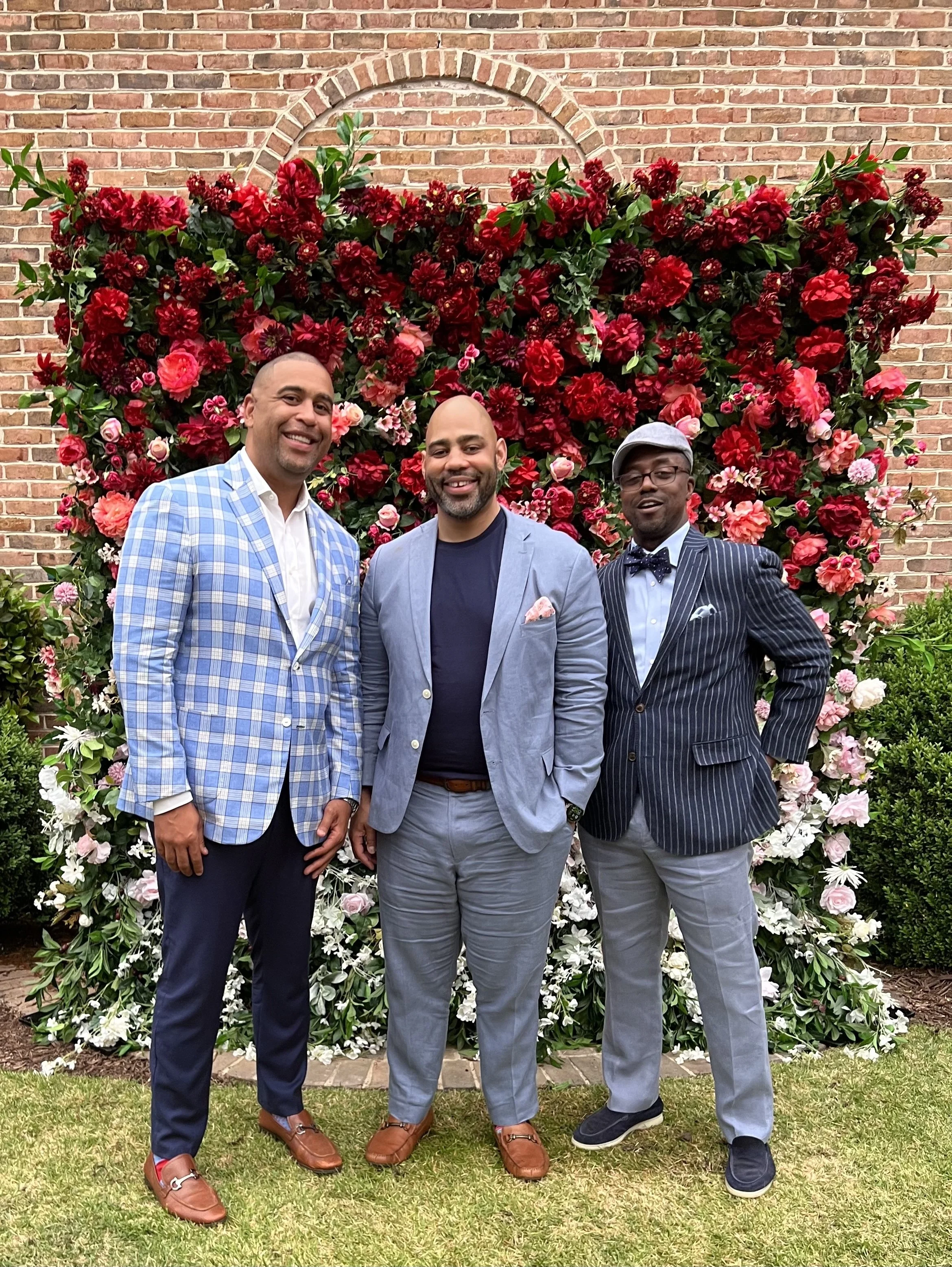 Three men in suits and smart attire standing in front of a flower wall with red, pink, and white flowers and a brick wall background.