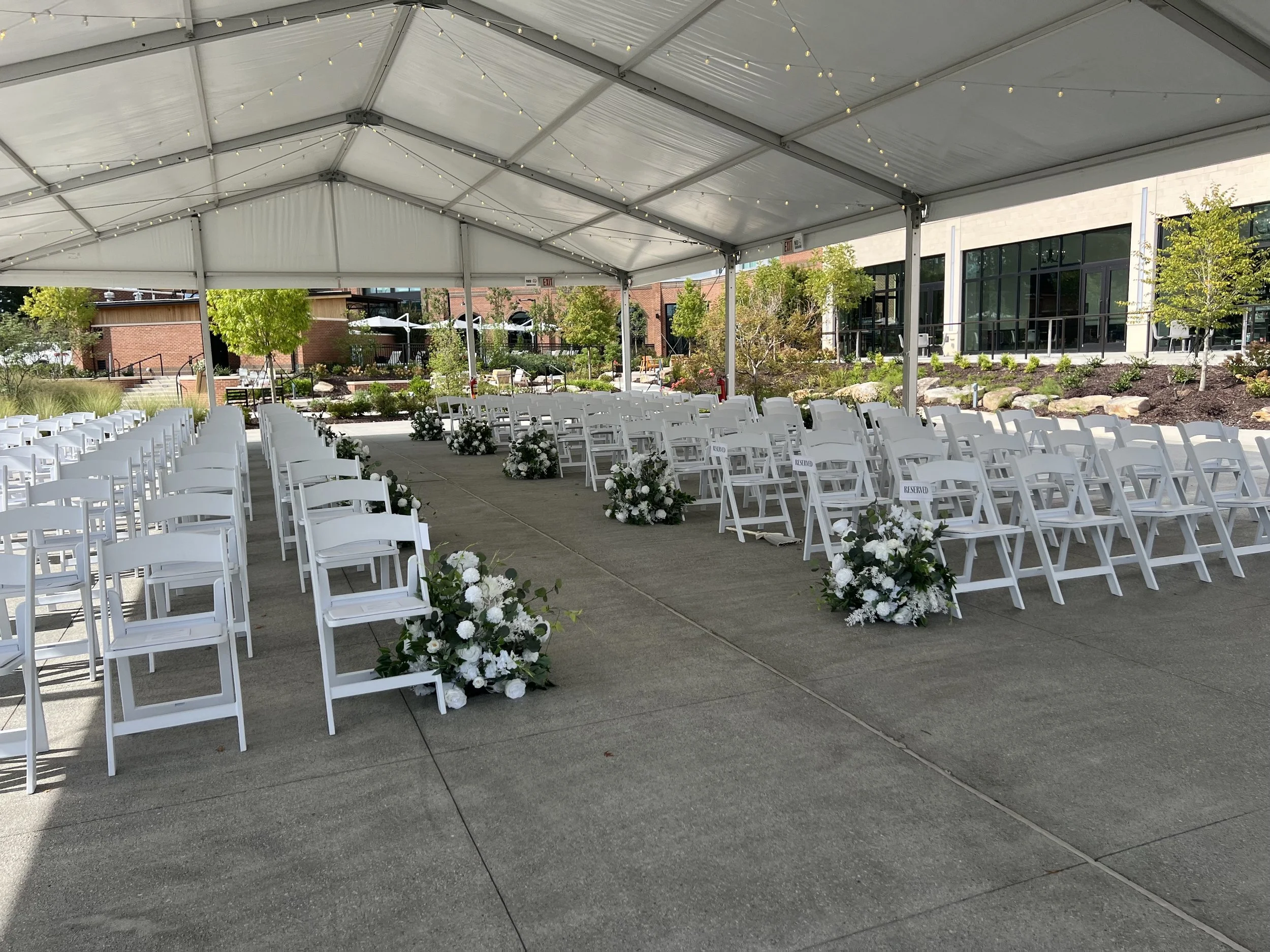 Outdoor wedding ceremony setup under a large white tent with white chairs arranged in rows, decorated with white and green floral arrangements, on a concrete surface surrounded by trees and buildings.