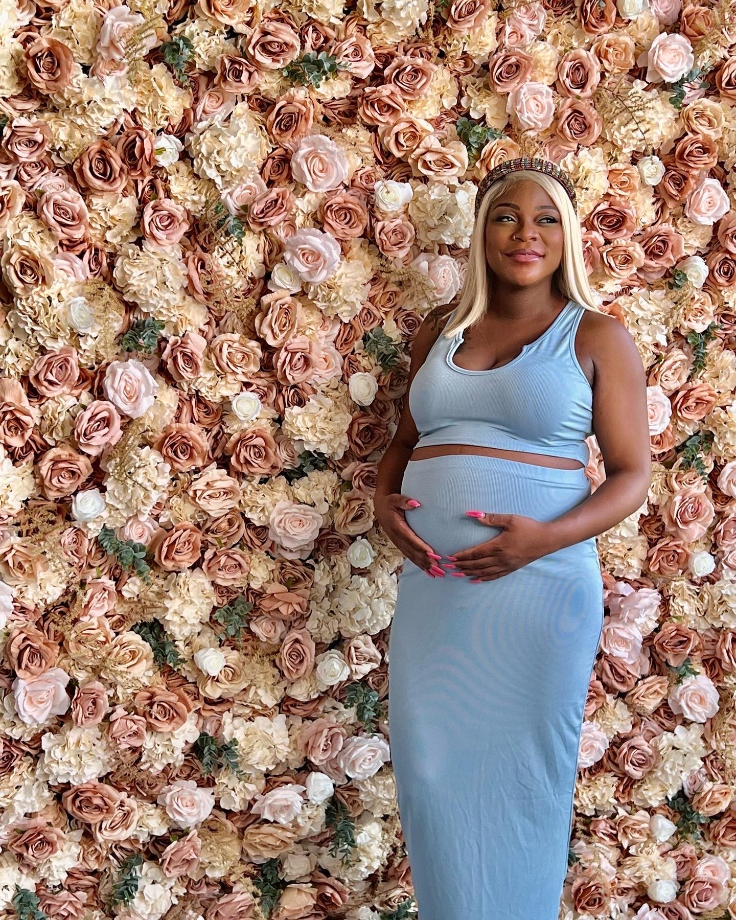 A pregnant woman in a blue outfit with a floral headband standing in front of a wall full of pink and white roses and hydrangeas, gently holding her belly and smiling.